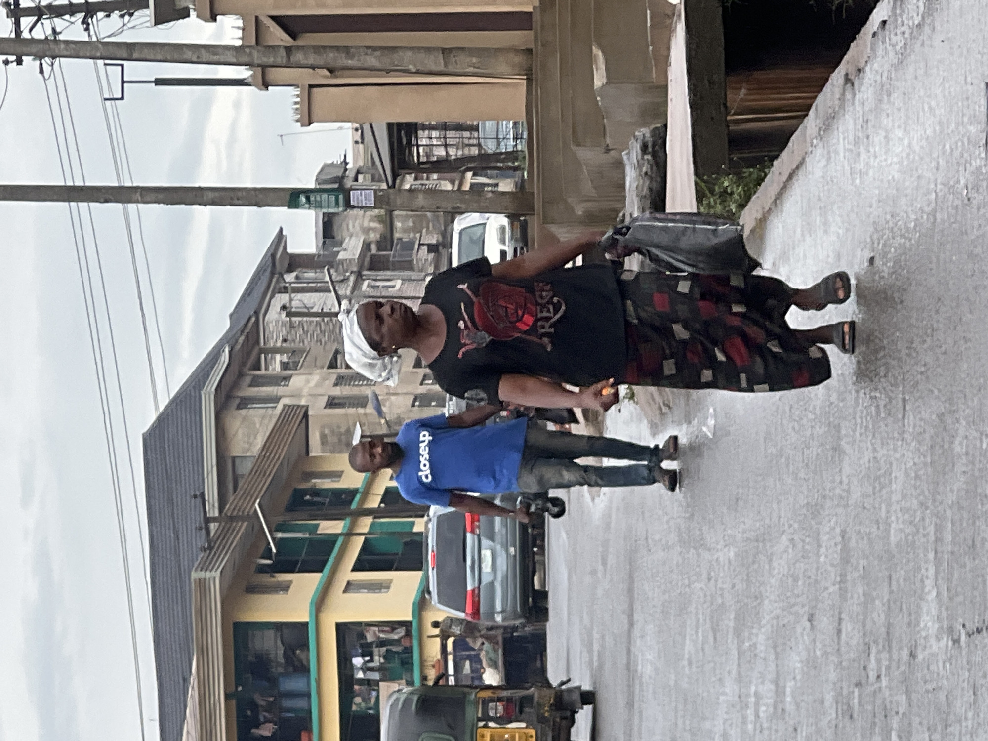 The image captures a street scene in Nkpor, Nigeria. Two individuals are walking on a wet, paved road.
The person in the foreground, on the left, is a man wearing a bright blue t-shirt with white text that reads "closeup" and dark pants. He is looking towards the viewer. He is carrying a black object in his right hand, possibly a bag or a tool.
To his right, a woman is walking away from the camera. She is wearing a black t-shirt with a graphic design and a patterned skirt with red and black squares. She has a white head covering. She is carrying a large black bag in her left hand.
In the background, there are multi-story buildings with residential and commercial spaces. Some of the buildings have balconies and various architectural styles. A grey car and a three-wheeled vehicle (a trishaw or Keke Napep) are visible on the road. The road surface appears wet, suggesting it might have rained recently or is currently drizzling. The sky is overcast, indicating a cloudy day. The overall atmosphere is that of a typical urban environment in Nigeria.