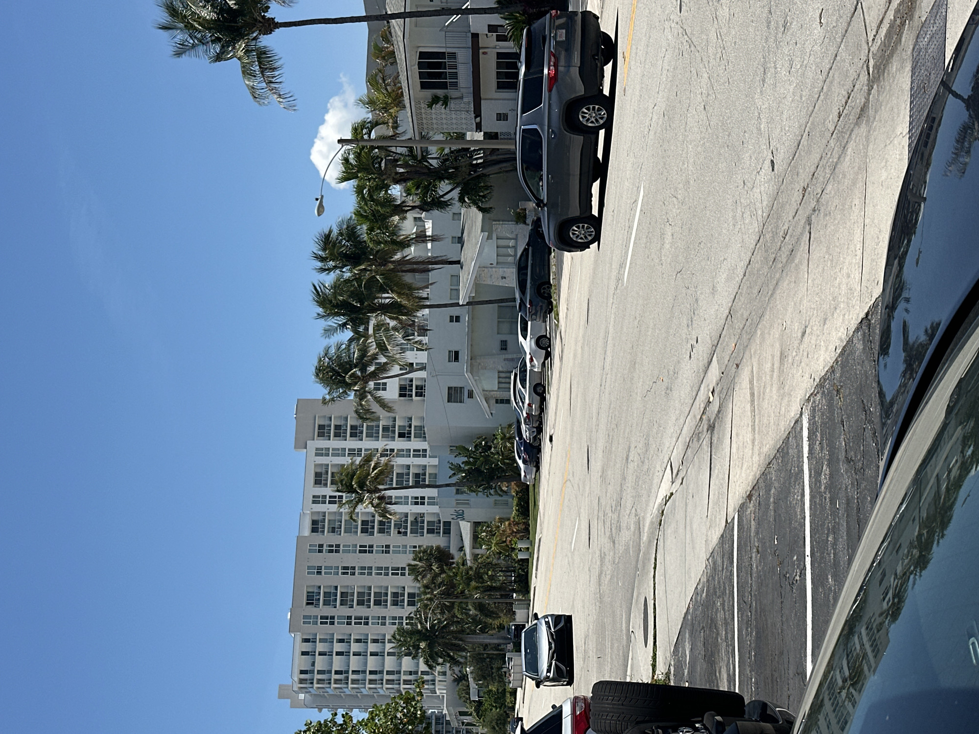 This photograph depicts a street scene in Fort Lauderdale, Florida, United States of America. The view is taken from within a vehicle, with a portion of the car's interior visible in the foreground. Outside, a line of parked cars stretches along the right side of the street. Palm trees are interspersed with white, high-rise buildings lining the opposite side of the street. The sky is clear and blue.