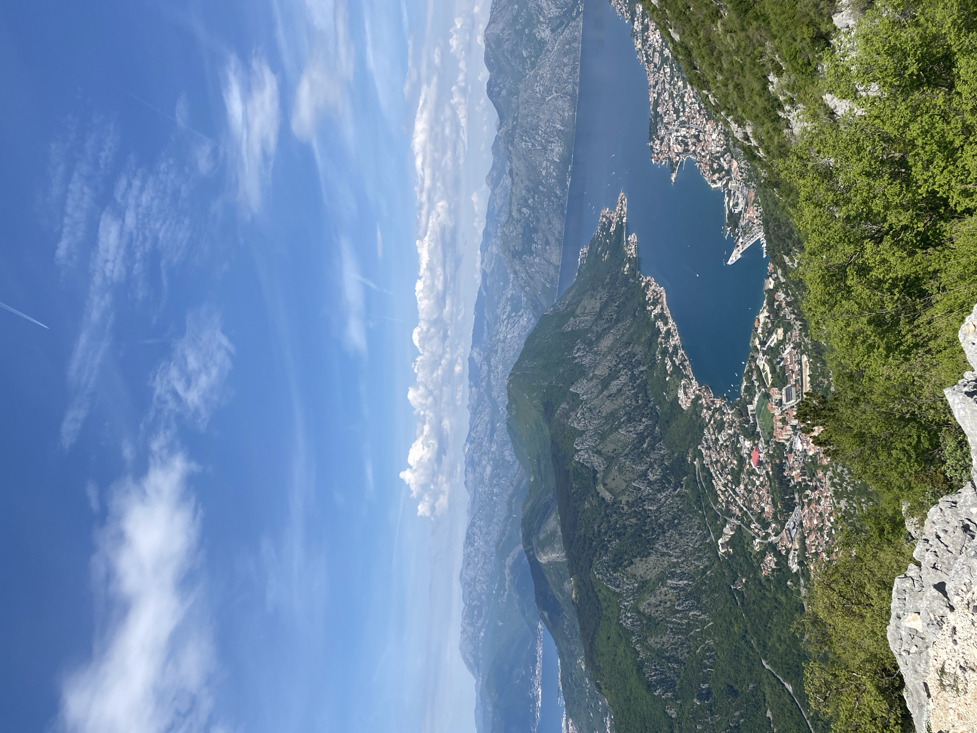 This elevated panoramic shot captures a stunning mountainous coastal landscape under a bright, clear blue sky. The viewpoint is from a high vantage point, likely a mountain ridge or cliff, looking down and across a vast expanse.
The upper portion of the image is dominated by the vibrant blue sky, adorned with scattered white clouds, ranging from wispy cirrus to more defined cumulus formations. Several faint, linear contrails from aircraft are visible stretching across the sky.
Below the sky, successive layers of majestic mountains recede into the distance, their rugged peaks and slopes covered in a mix of lush green vegetation and exposed grey rock faces. The lighting suggests strong midday sun, illuminating the landscape and casting subtle shadows that highlight the topographical relief.
A prominent feature is a large, deep blue body of water, resembling a bay or fjord, that winds through the mountainous terrain on the right side of the frame. This body of water forms a distinctive, almost heart-shaped inlet in the mid-right, within which a densely packed town is nestled. The town features numerous buildings with red-tiled roofs, extending along the shoreline and climbing up the lower slopes of the surrounding mountains. Small white boats dot the calm surface of the water, particularly within the heart-shaped inlet, indicating a bustling harbor or recreational area.
The immediate foreground on the right consists of steep, rocky terrain covered in vibrant green foliage, emphasizing the considerable height from which the photograph was taken. The scene is devoid of any specific human activity in detail or visible text.
The dramatic scenery, with its towering mountains, serene blue water, and picturesque town, suggests a location of significant natural beauty. Given the location context of Žanjev Do, Montenegro, this breathtaking view is of the renowned Bay of Kotor, with the historic town of Kotor visible at its innermost part.