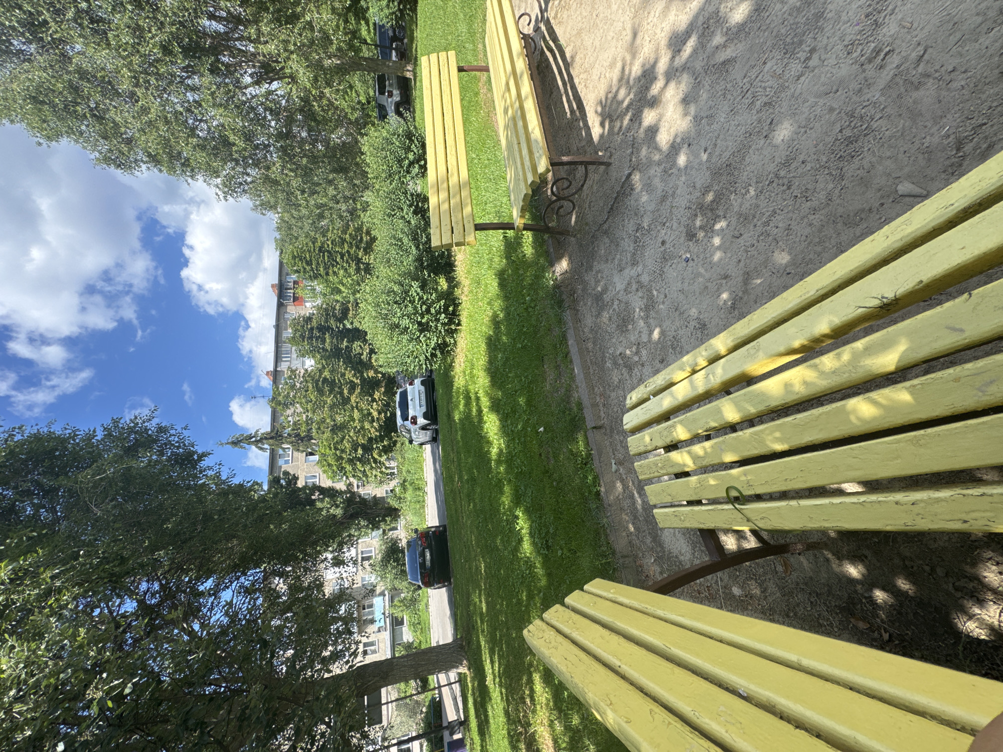 The image depicts a park scene in Poltava, Ukraine, on a sunny day with scattered clouds. In the foreground, two weathered yellow wooden benches are positioned on a gravel path. Behind the benches, a well-maintained lawn stretches towards a paved road where two cars are parked: a dark sedan and a white hatchback. Beyond the road, a dense line of green trees and bushes forms a backdrop. In the distance, a multi-story apartment building with a light-colored facade and reddish-orange rooflines is visible. The overall environment suggests a residential area with green spaces.