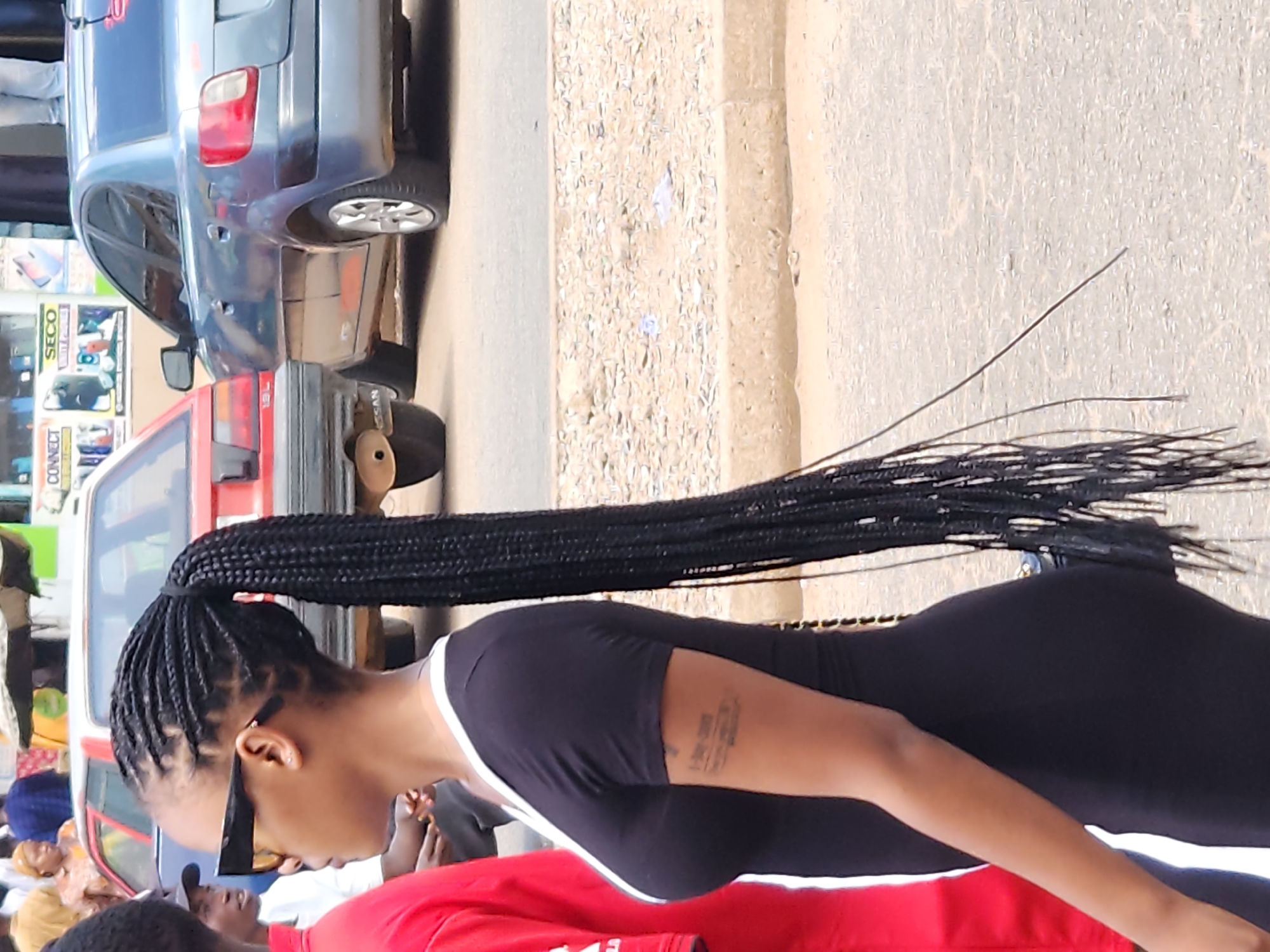 A person with long, braided hair is standing outside, wearing a black dress with a white stripe. They are on a street next to a red car and a blue car. The environment appears to be a city street, with other people visible in the background.