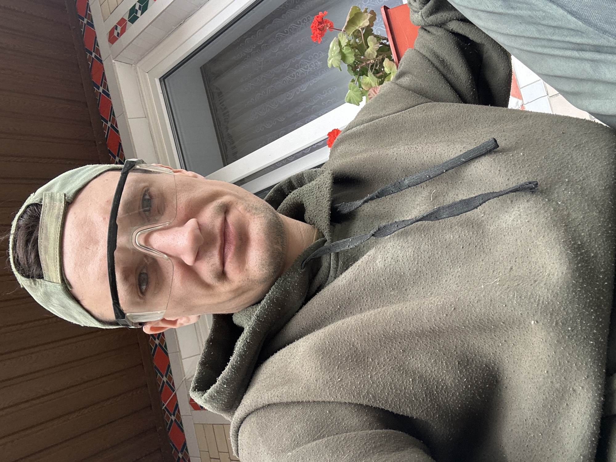In Makiv, Ukraine, a selfie shows a man wearing a camouflage baseball cap, clear protective glasses, and a faded olive green hoodie with pilling. He is positioned outdoors, under a brown wooden overhang with decorative tile trim. To his right, a window with sheer curtains is visible, and in front of the window, a red flower pot holds red geraniums. The man's left shoulder and arm are visible, as well as a portion of his right leg, which appears to be clad in grey pants. The overall impression is that of an individual engaged in some form of outdoor activity or work.
