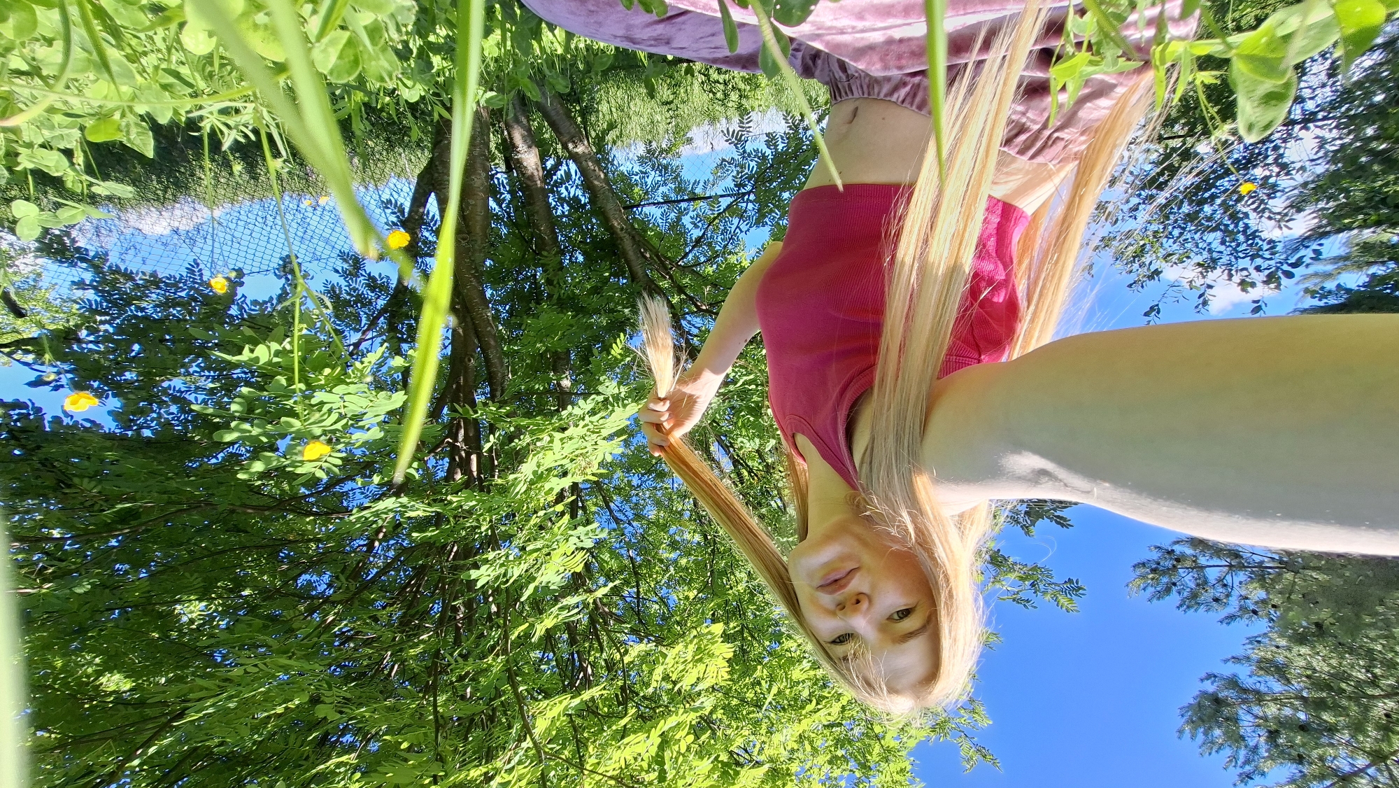 A young Caucasian woman with long blonde hair is taking a selfie in a grassy outdoor area. She is wearing a pink tank top and light pink velvet pants. She is holding a section of her hair in her right hand and looking towards the camera. The background features lush green trees with bright green leaves, yellow wildflowers, and a clear blue sky. The image appears to be taken on a sunny day, possibly in a park or natural setting. There are no visible structures or specific landmarks that would indicate a precise location within Bolshoy Bukor, Russia. The environment is characterized by abundant vegetation and natural light.