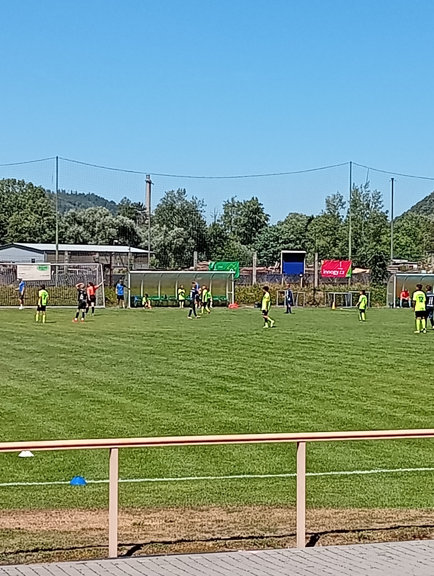 The image depicts a junior football (soccer) match taking place on a grass field under a clear blue sky. Several young players, mostly dressed in yellow and black or yellow and white uniforms, are visible on the field. Some are actively playing, while others are positioned on the sidelines or in front of a dugout. In the background, there are trees, a fence, and some buildings. A prominent banner with the text "Innogy cz" is displayed on the fence. The scene appears to be a sports facility located in Tišnov, Czechia.