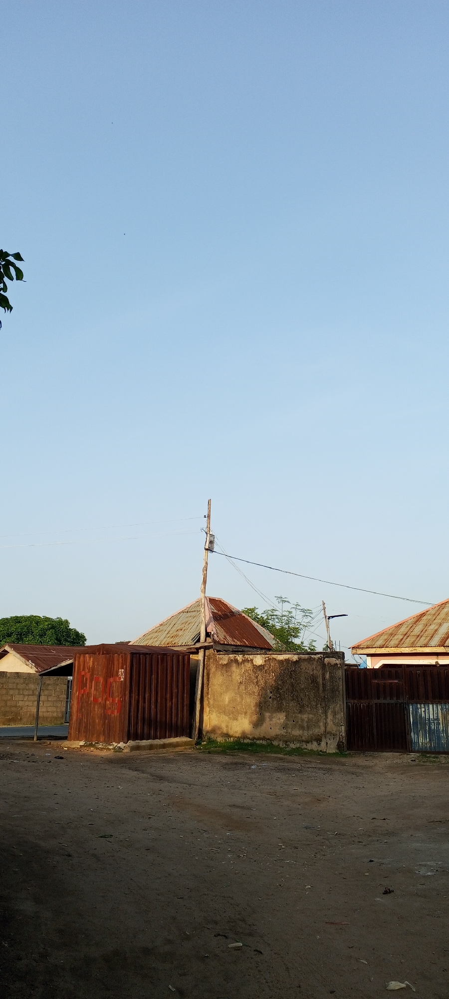 The image depicts an outdoor scene in a dusty urban environment, likely in Jalingo, Nigeria, given the architectural style and the presence of corrugated metal roofing, which is common in the region. The time of day appears to be late afternoon or early morning, with soft, diffused sunlight illuminating the scene.
In the foreground, a dirt road is visible, characterized by loose soil and scattered debris. To the left of the road, a rusty corrugated metal fence stretches across the frame, partially obscuring the view beyond. Behind this fence, a section of a building with a similar corrugated metal roof is visible, showing signs of age and weathering.
Further into the image, a concrete wall with a rough texture runs along the right side. A wooden utility pole stands adjacent to this wall, supporting electrical wires and a small electrical box. Beyond the wall, more corrugated metal roofs of different structures can be seen, some appearing to be residences or small businesses. The roofs are a mix of oxidized metal and what might be painted surfaces.
The background consists of a clear, pale blue sky with a few faint specks that could be distant birds or atmospheric particles. A tree with green foliage is partially visible on the far left edge of the frame, suggesting the presence of vegetation in the vicinity. Another cluster of trees with finer branches is seen behind the utility pole and between some of the buildings.
No people are visible in the image, and there are no apparent ongoing actions or interactions. The overall impression is one of a somewhat utilitarian and possibly developing urban area, with an emphasis on functional, weather-resistant building materials. The scene is static and devoid of any specific human activity.