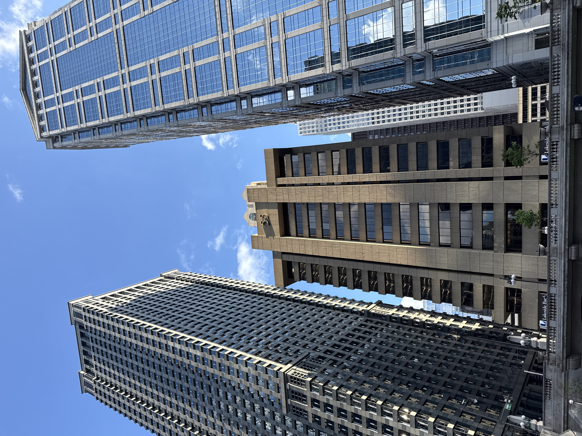The image captures a low-angle view of several skyscrapers in Chicago, United States of America, under a blue sky with scattered clouds. The buildings have different architectural styles, including a modern glass-clad tower, a stone-faced building with recessed windows, and a dark, grid-like structure. The perspective emphasizes the height and scale of the buildings, creating a dynamic composition. A portion of a bridge or elevated walkway is visible on the right side. The reflections on the glass windows indicate the presence of other buildings and the sky. A Lakeside Bank sign is visible on one of the buildings.