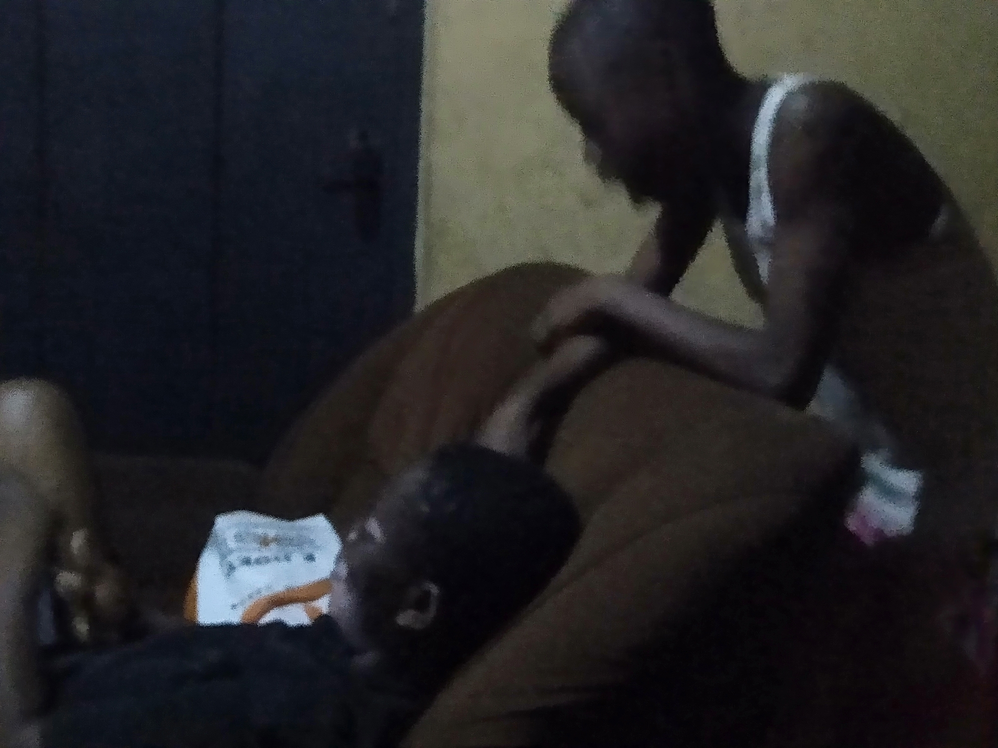 Two people are in a dimly lit room. One person is lying on a brown couch, and the other is standing and leaning over the couch. There is a package with white, orange, and grey colors next to the person lying down. A dark door is partially visible in the background on the left side of the image. The background wall has a yellow hue. The location is Shangisha, Nigeria.