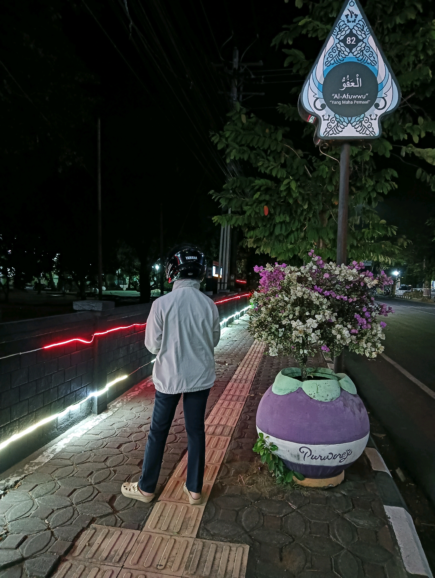 The image depicts a nighttime scene on a sidewalk in Purworejo, Indonesia. A person wearing a white jacket, dark pants, and a black helmet stands facing away from the camera, looking towards a decorative sign and a flowering plant. The sign, shaped like a traditional Javanese "pusaka" (dagger), features Arabic calligraphy and the text "AL-AFUUWWU" and "Yang Maha Pemaaf" (The All-Forgiving). A potted plant with pink and white flowers sits next to the sign. The sidewalk is illuminated by red and white LED lights embedded in a brick wall. The street is visible to the right, and trees and other foliage are present in the background. A large, round, purple planter with a white band inscribed with "Purworejo" is visible in the foreground.