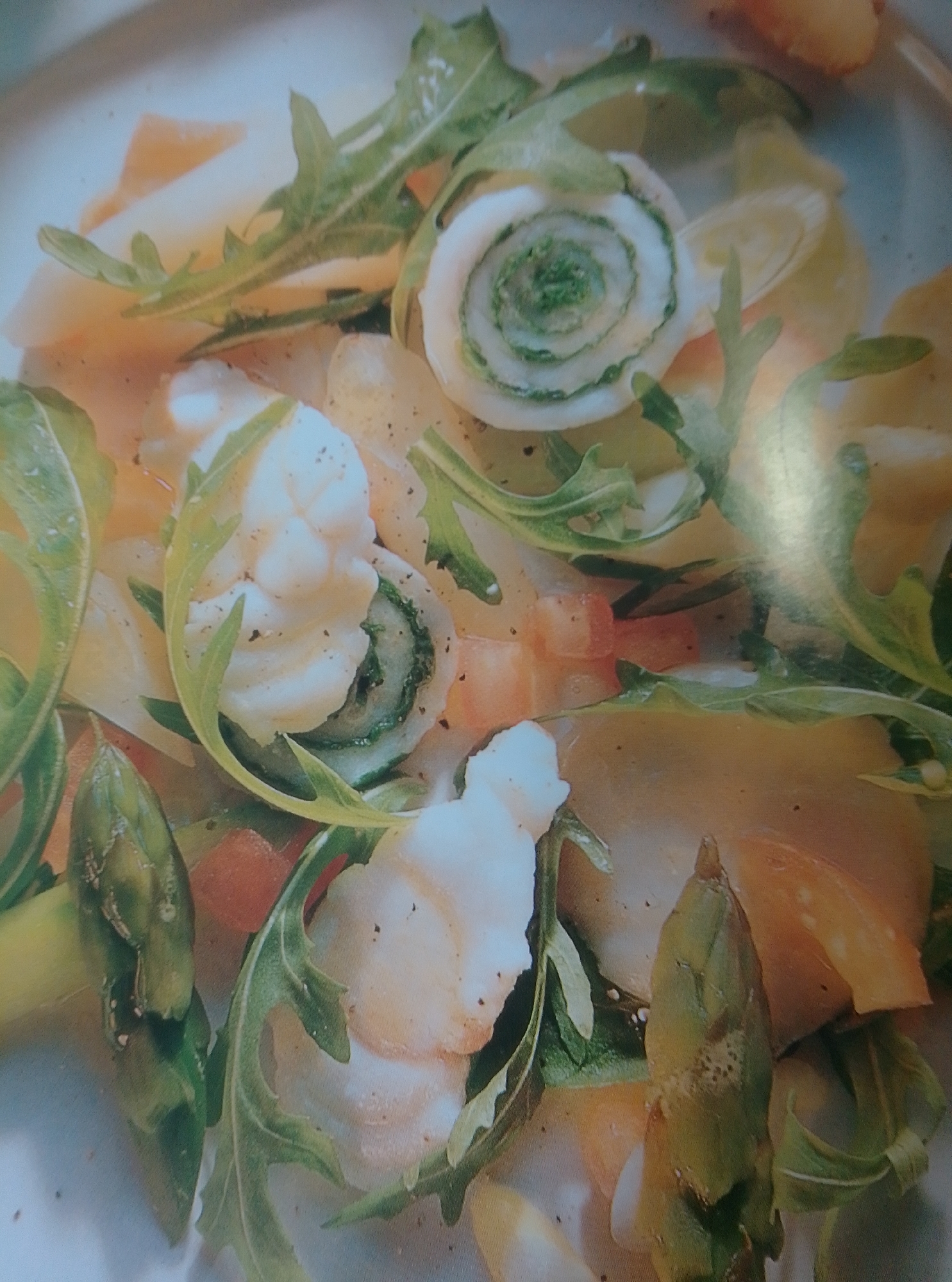 A close-up, overhead shot captures a gourmet seafood salad. The dish features white seafood pieces, possibly shrimp or scallops, arranged amongst pale, thinly sliced vegetables and vibrant green arugula. A distinctive spiral-shaped element, appearing to be a rolled slice of cucumber with green filling, is prominent in the center. The salad is seasoned with black pepper and appears to be served in a light-colored bowl or on a plate. The lighting creates a soft, slightly blurred effect, emphasizing the fresh textures and colors of the ingredients. This image is not located in the city of Wedel, Germany.
