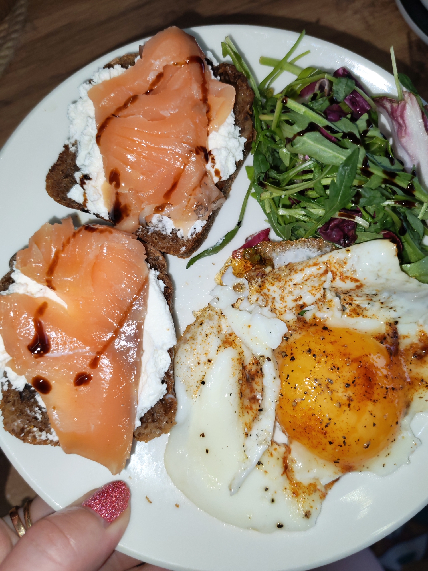 A top-down shot captures a freshly prepared meal, likely breakfast or brunch, served on a white round plate. The composition suggests an indoor setting, possibly in Izhevsk, Russia.
On the left side of the plate, there are two small, roughly square slices of dark bread, which appear to be rye or whole grain. Each slice is topped with a generous spread of white, creamy cheese, resembling cottage cheese or ricotta. Laid over the cheese are several delicate, pink slices of smoked salmon, artfully drizzled with a dark, glossy sauce, likely balsamic glaze.
Adjacent to the salmon toasts is a fresh green salad, composed primarily of arugula and hints of reddish-purple radicchio, also lightly dressed with a dark vinaigrette. Below the salad and next to the toasts is a perfectly cooked fried egg. It features a bright, round, runny orange yolk surrounded by a crispy-edged white, seasoned generously with black pepper and a reddish spice, possibly paprika.
A hand, adorned with sparkling red nail polish and two gold rings on the ring finger, holds the edge of the plate, suggesting the meal is ready to be eaten. The background shows a warm, wooden surface, likely a table. The lighting is artificial and warm, illuminating the food. There is no visible text.