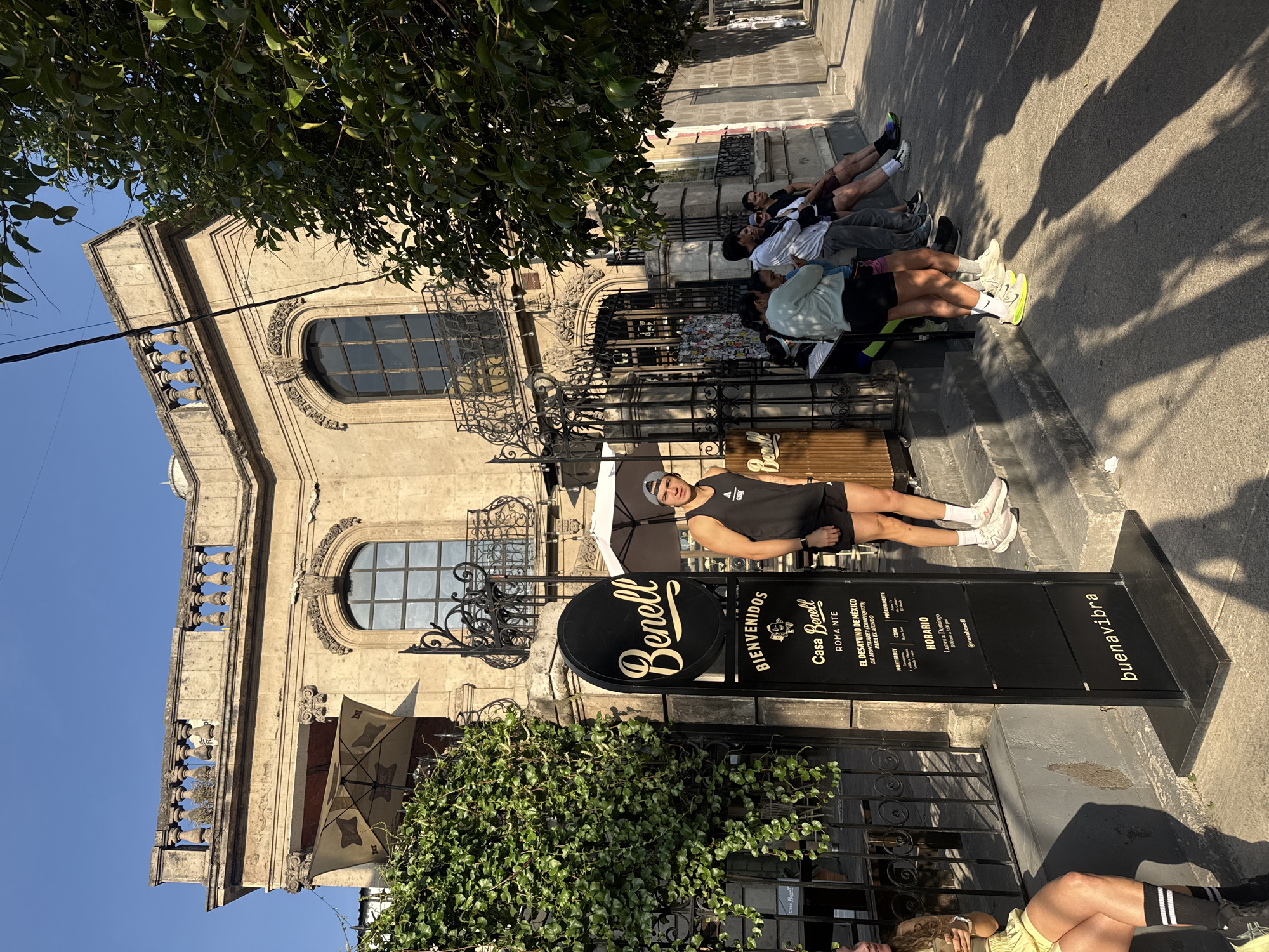 A vibrant daytime street scene in Mexico City, Mexico, depicts several individuals relaxing on a sidewalk outside a cafe. The background features a grand, multi-story building with ornate architecture, including arched windows with decorative grilles and balconies with balustrades. Lush green foliage from an overhead tree and ivy growing on the building adds to the natural elements of the setting.
In the foreground, a man in a black tank top, black shorts, a grey baseball cap, and white sneakers lies on his back, propped up on his elbows, looking towards the camera. Further along the sidewalk, other casually dressed people are seated or lounging, some with their legs stretched out. Strong shadows cast across the sidewalk from the people and nearby trees indicate bright, sunny weather, likely in the morning or late afternoon.
Prominently displayed are two black signs for "Casa Benell". A circular sign features "Benell" in white script. A rectangular sign next to it reads "BIENVENIDOS", "Casa Benell", "ROMA NTE", and details about "EL DESAYUNO DE MÉXICO DE MONTERREY (TAMPIQUITO) PARA EL MUNDO". It also lists "HORARIO Lunes a Domingo 8:00 am a 1:00 pm" and mentions "@casabenell" and "buenavibra". Other text includes "MONTERREY", "CONX", "PROXIMAMENTE", and "GISK". The "ROMA NTE" (Roma Norte) reference helps confirm the Mexico City location. The people appear to be taking a break or waiting, exuding a relaxed atmosphere.