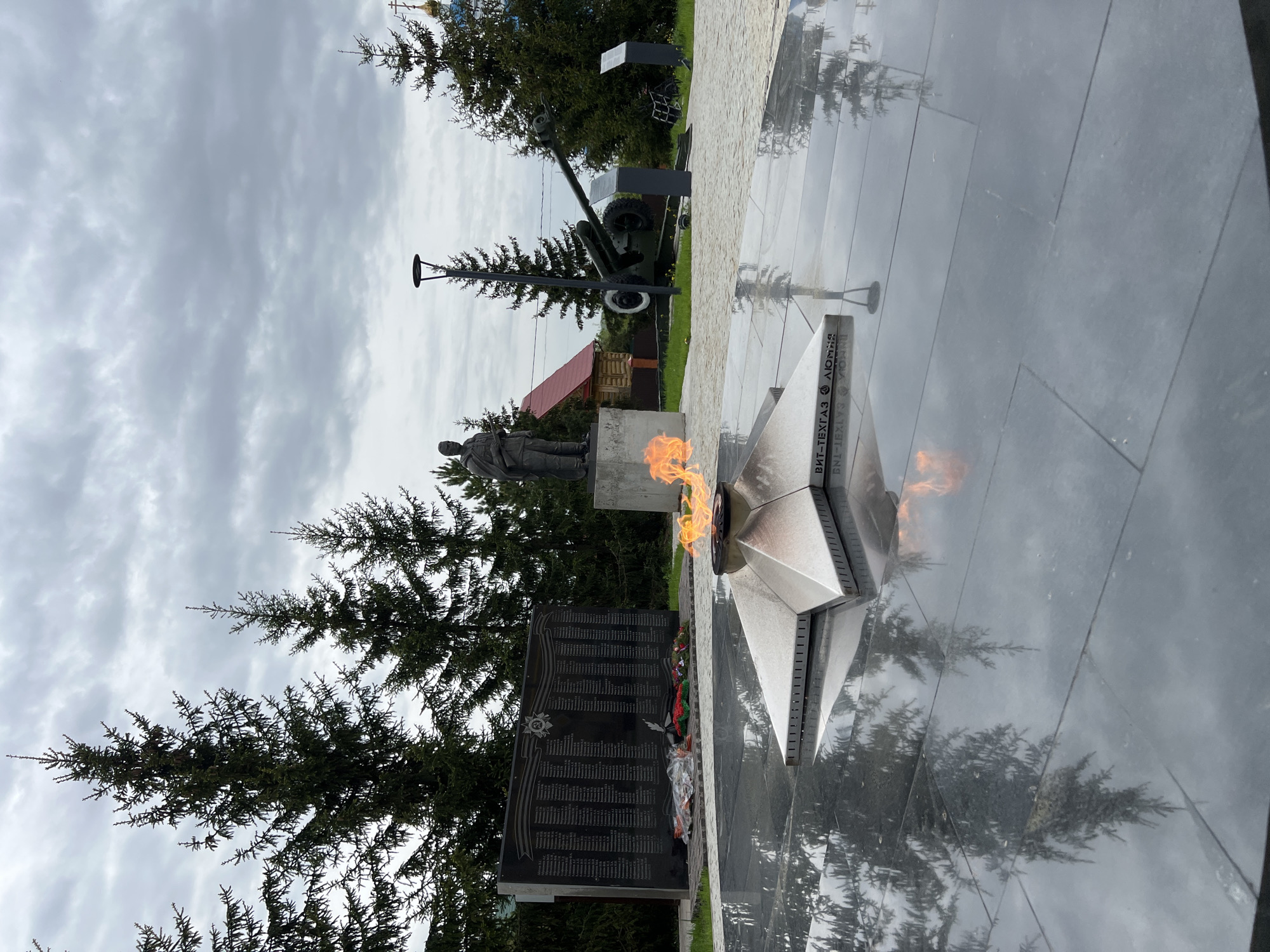 The image shows a war memorial in Введенская Слобода, Russia. The memorial features an eternal flame, a statue of a soldier, and a black granite wall inscribed with names. A cannon is also present. The ground around the memorial is dark and reflective, with trees in the background and a cloudy sky overhead.