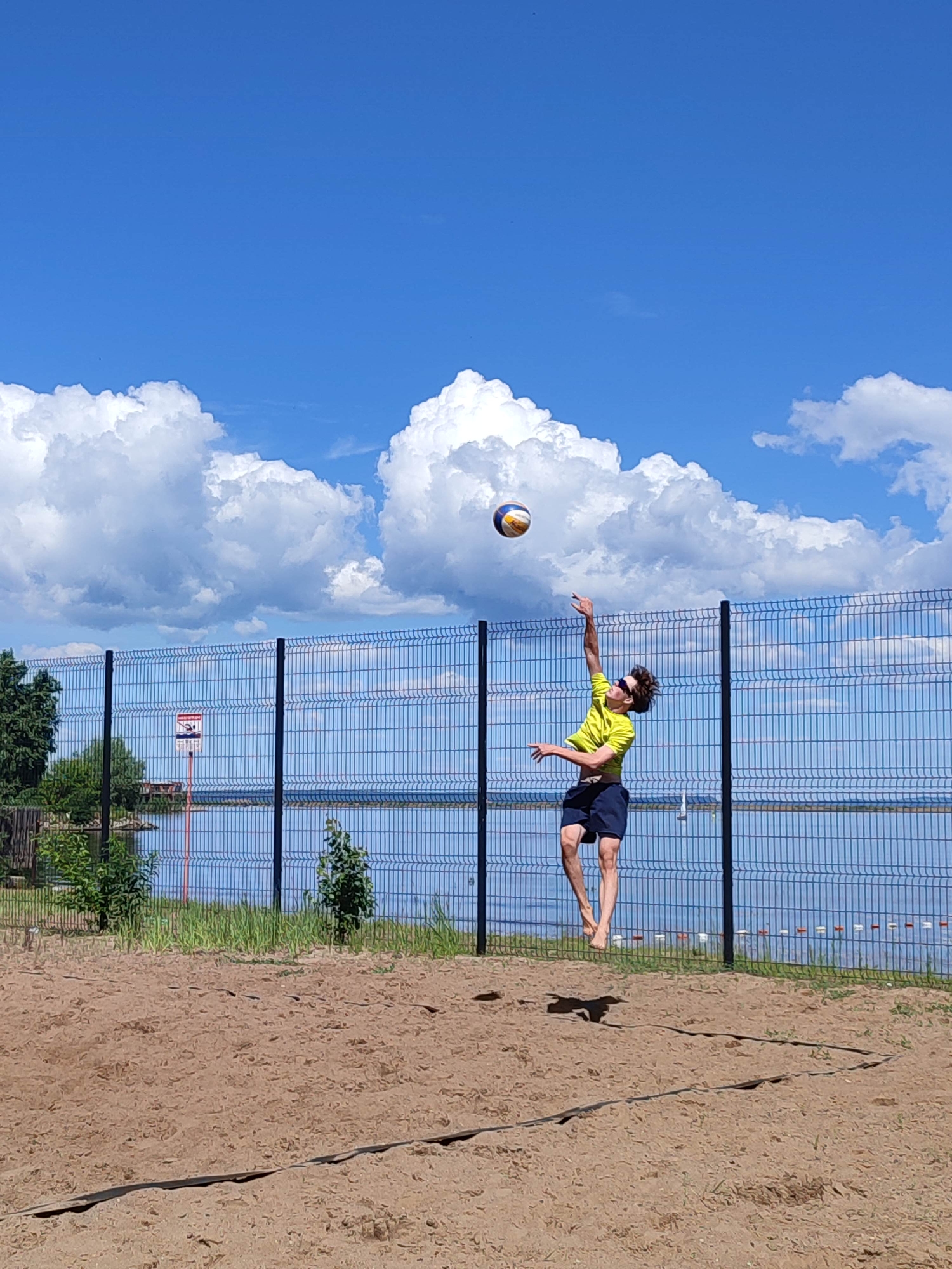 The image depicts an outdoor beach volleyball court on a sunny day. A single male individual is captured mid-air, performing a volleyball play. He is positioned in the middle ground, slightly right of the frame's vertical center. He wears a bright yellow short-sleeved t-shirt, dark blue shorts, and sunglasses, and is barefoot. His right arm is extended upward, bent at the elbow, hand open, directed towards a multi-colored (blue, yellow, white, brown) volleyball situated above and slightly to his left. His left arm is bent and held near his chest. His legs are bent at the knees, and he casts a prominent shadow on the sand below him.
The foreground consists of a light brown sandy court, with dark boundary lines visible on the right. Behind the player, a dark metal mesh fence, characterized by vertical posts and a grid pattern, spans horizontally across the scene. Beyond this fence lies a calm, blue body of water, likely a lake or wide river, reflecting the sky. Along the water's edge, sparse green vegetation, including small trees and bushes, is visible. On the far left, through the fence and above the water, a red and white sign mounted on a red pole is partially discernible. The upper portion of the image is dominated by a clear blue sky dotted with several large, white cumulus clouds. The lighting is bright, indicative of direct sunlight. The location is an unspecified outdoor recreational area within an unknown city.