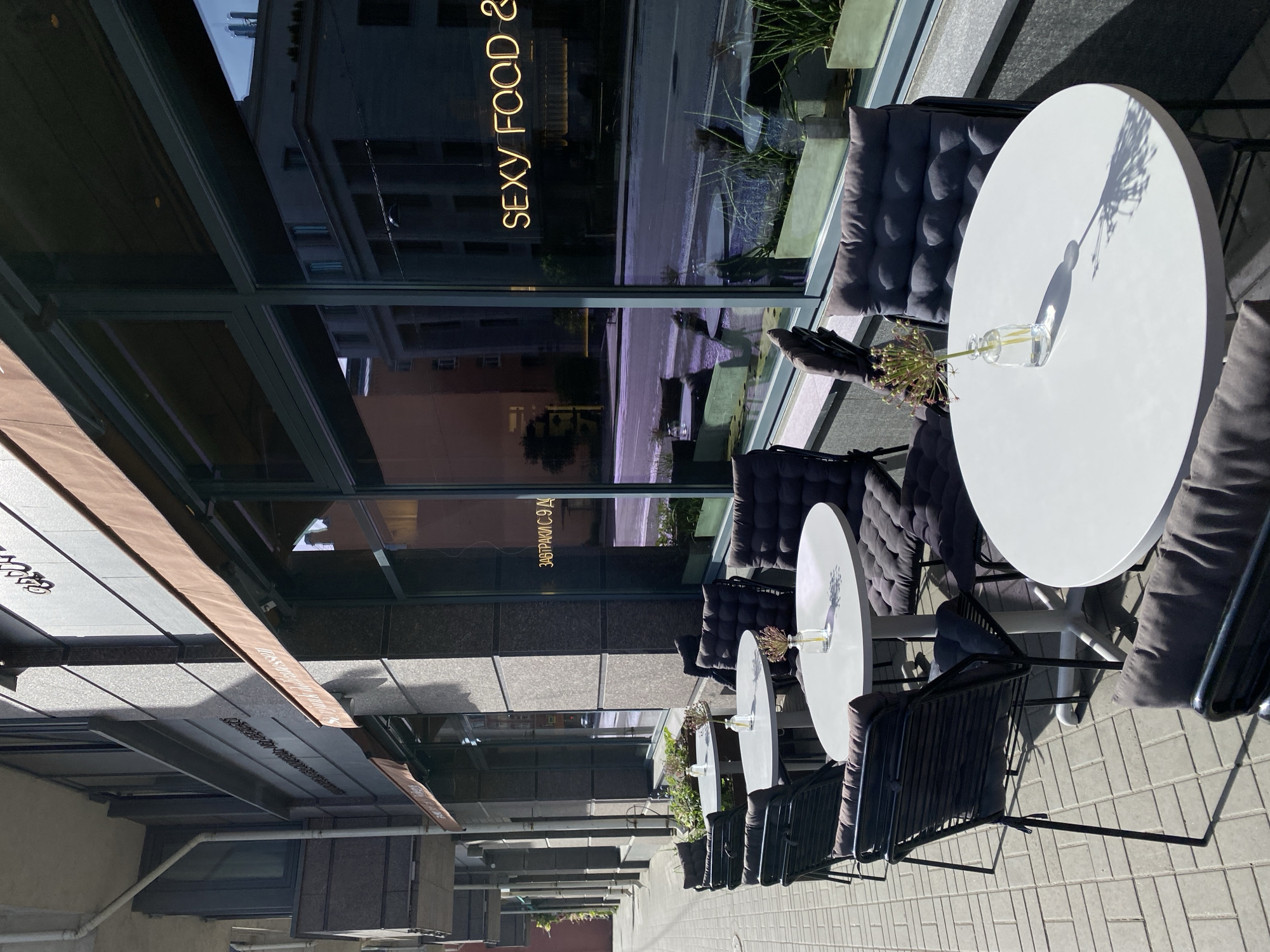 The image depicts an outdoor seating area of a restaurant or cafe in Saint Petersburg, Russia. A row of round, white tables with black chairs and gray cushions lines the exterior of a building with large windows. Each table features a small glass vase with flowers. The windows reflect the street scene and buildings across the street, including signage visible through the glass that reads "SEXY FOOD &". The scene suggests a modern, urban environment with elements of outdoor dining and reflection of the cityscape. The environment appears to be a sunny, daytime setting.