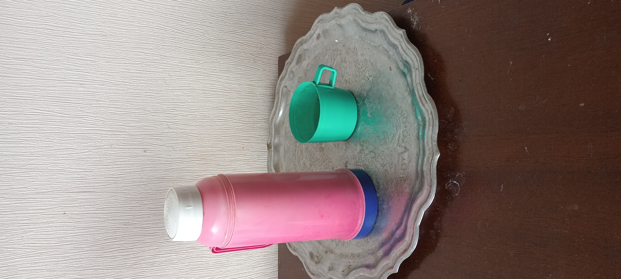 The image shows a pink thermos with a white lid and a blue base, positioned on the left side of a decorative silver tray. To the right of the thermos, also on the tray, is a green cup with a handle. The tray itself has an ornate, embossed pattern and is resting on a dark brown wooden surface, likely a table. The background is a textured, off-white wall. There are no people or animals visible in the image, and no actions or interactions are taking place. The scene is indoors, and the lighting appears to be natural or ambient, casting soft shadows. There is no indication of the specific location within Kyiv, Ukraine, as the image contains no recognizable landmarks or contextual clues.