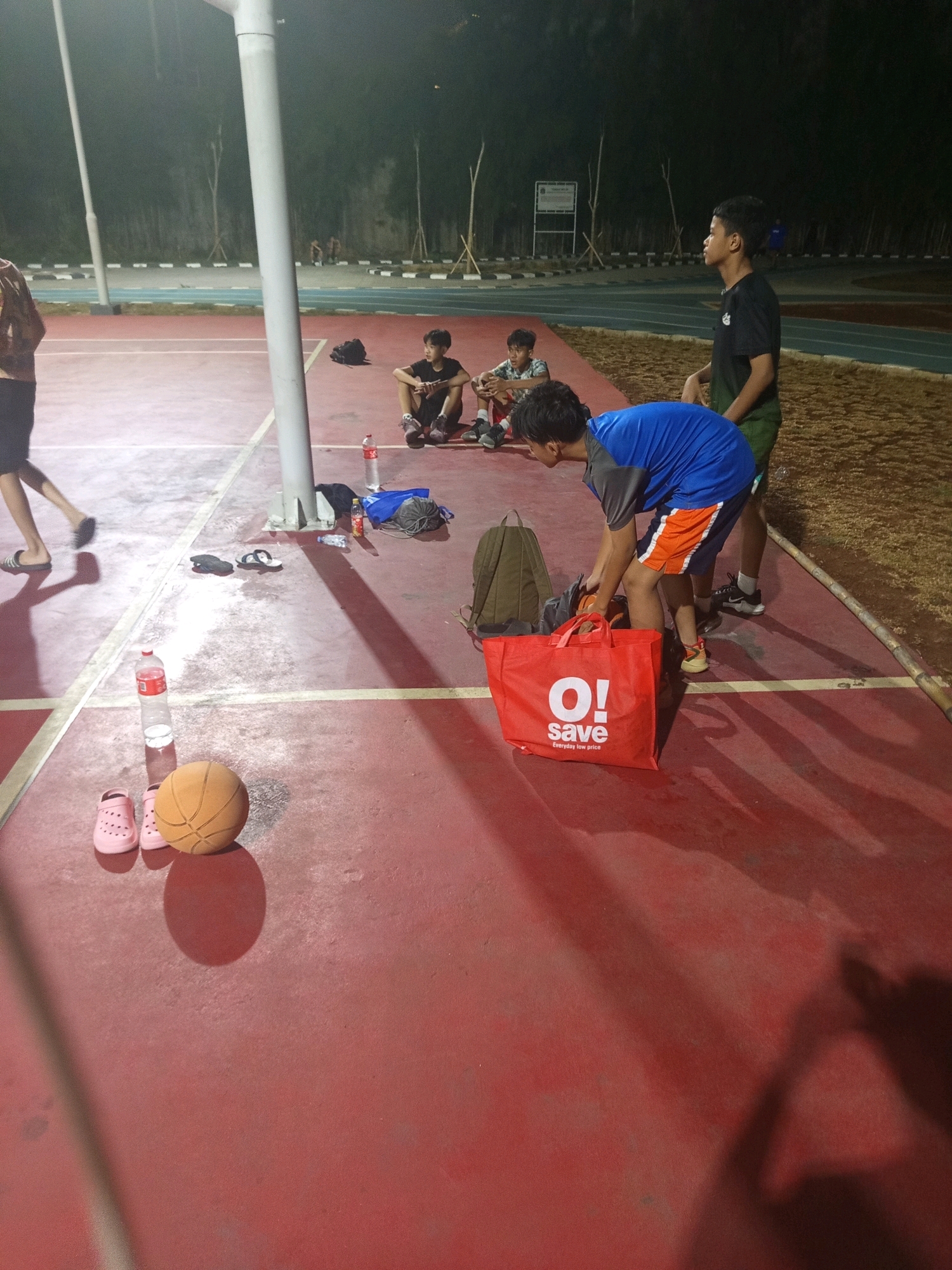 This is a low-angle, full shot of a group of young men on a red basketball court in West Jakarta, Indonesia, at night. A tall, white pole stands on the left side of the frame, casting a shadow. In the foreground, a basketball and a pair of pink clogs are on the court. A person in a blue shirt and orange shorts is bending down, holding a red shopping bag with "O! save" written on it. Two other young men are sitting on the court in the mid-ground. One is wearing a black shirt and green shorts and looking to the right. Another is wearing a patterned shirt and dark shorts and looking forward. In the background, a track and field facility is visible, with lights and trees in the distance. The lighting suggests it is nighttime, and the court appears to be well-lit.
