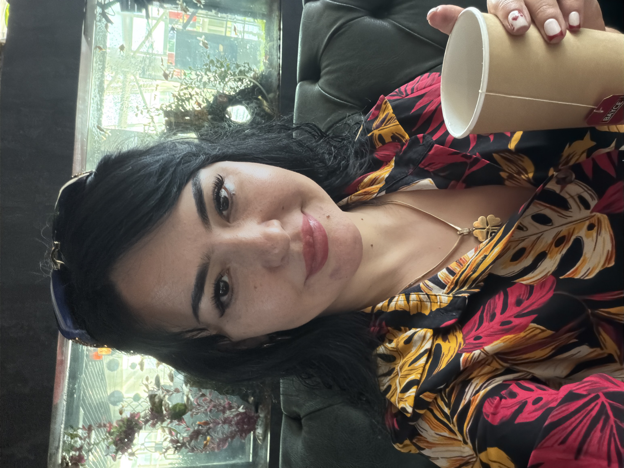 A woman with dark hair and fair skin is taking a selfie. She is wearing a colorful, patterned shirt and a necklace with a four-leaf clover pendant. She is holding a paper cup with a tea bag. Behind her is a dark green, tufted sofa and a large aquarium filled with small fish and artificial plants. Sunglasses are perched on her head. The environment suggests an indoor setting, possibly a cafe or restaurant, with a view into an aquarium. No specific landmarks of Samarkand City, Uzbekistan are visible.
