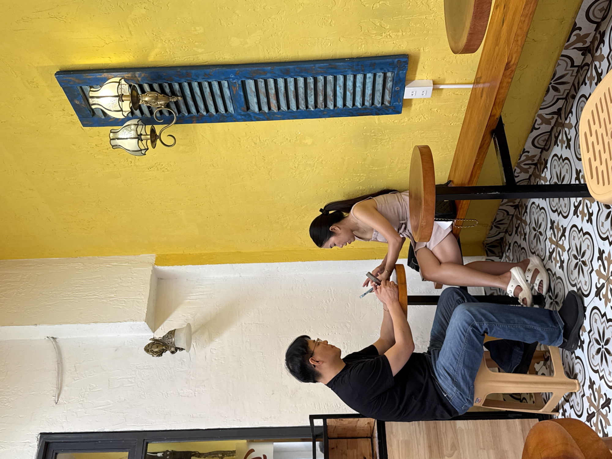 In Nha Trang Ward, Vietnam, an indoor scene features a man and a woman sitting at a table. The man, wearing a black t-shirt and blue jeans, is holding a smartphone. The woman, dressed in a light-colored top and shorts, also holds a smartphone. They appear to be interacting with their phones. The table they sit at has a round top, with wooden benches adjacent to them. The floor is tiled with a floral pattern. The wall behind the man is white, while the wall behind the woman is yellow, decorated with a blue louver and ornate wall-mounted lamps. The lighting is ambient. A window frame is visible at the bottom left.