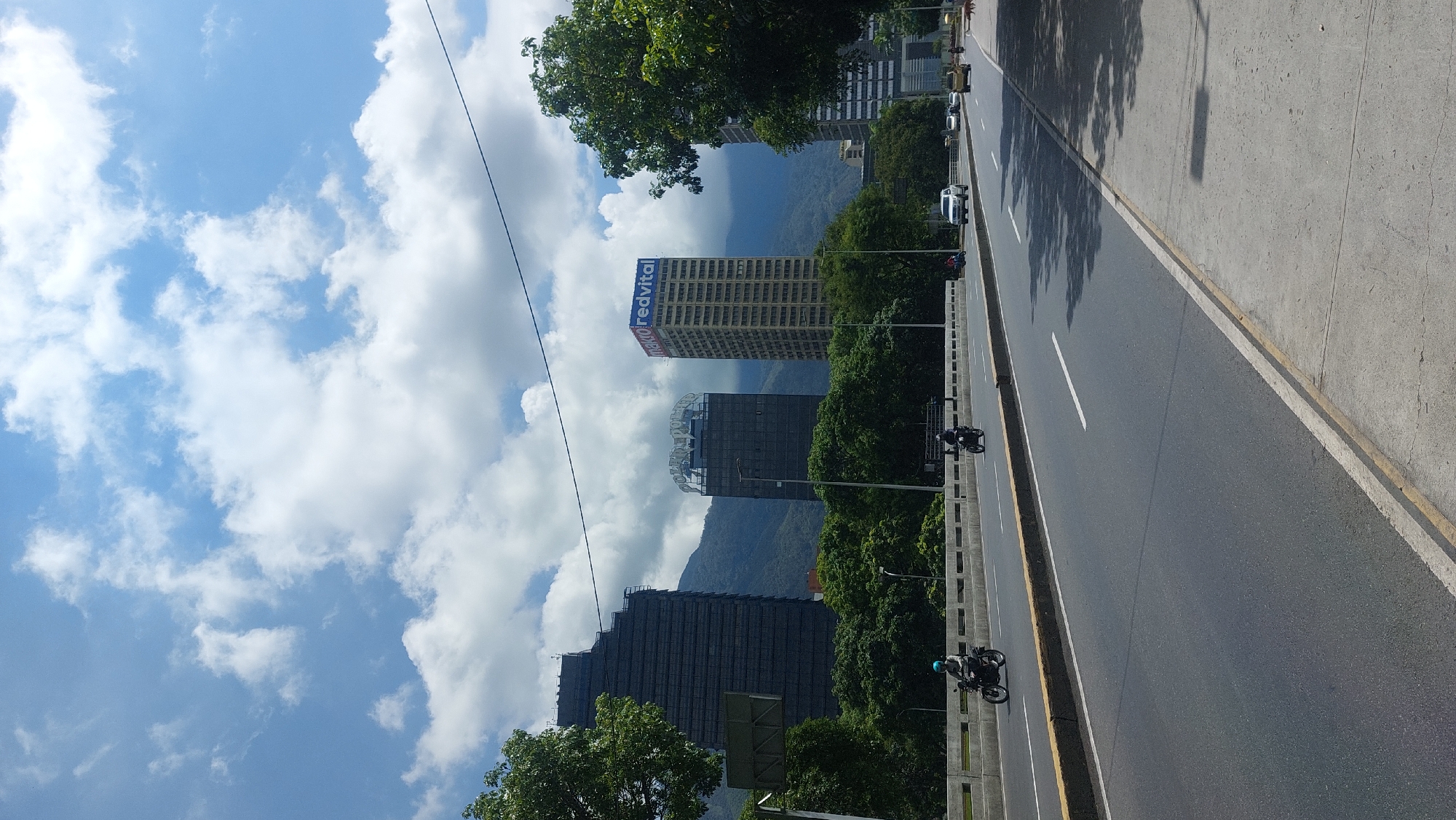 The image depicts a street scene in Caracas, Venezuela, showcasing modern buildings against a backdrop of mountains and a partly cloudy sky. The "redvital" building is visible among the high-rise structures. A multi-lane road runs alongside a green space, with two motorcycles moving in the same direction. The road is lined with concrete barriers, and the scene is captured on a sunny day.