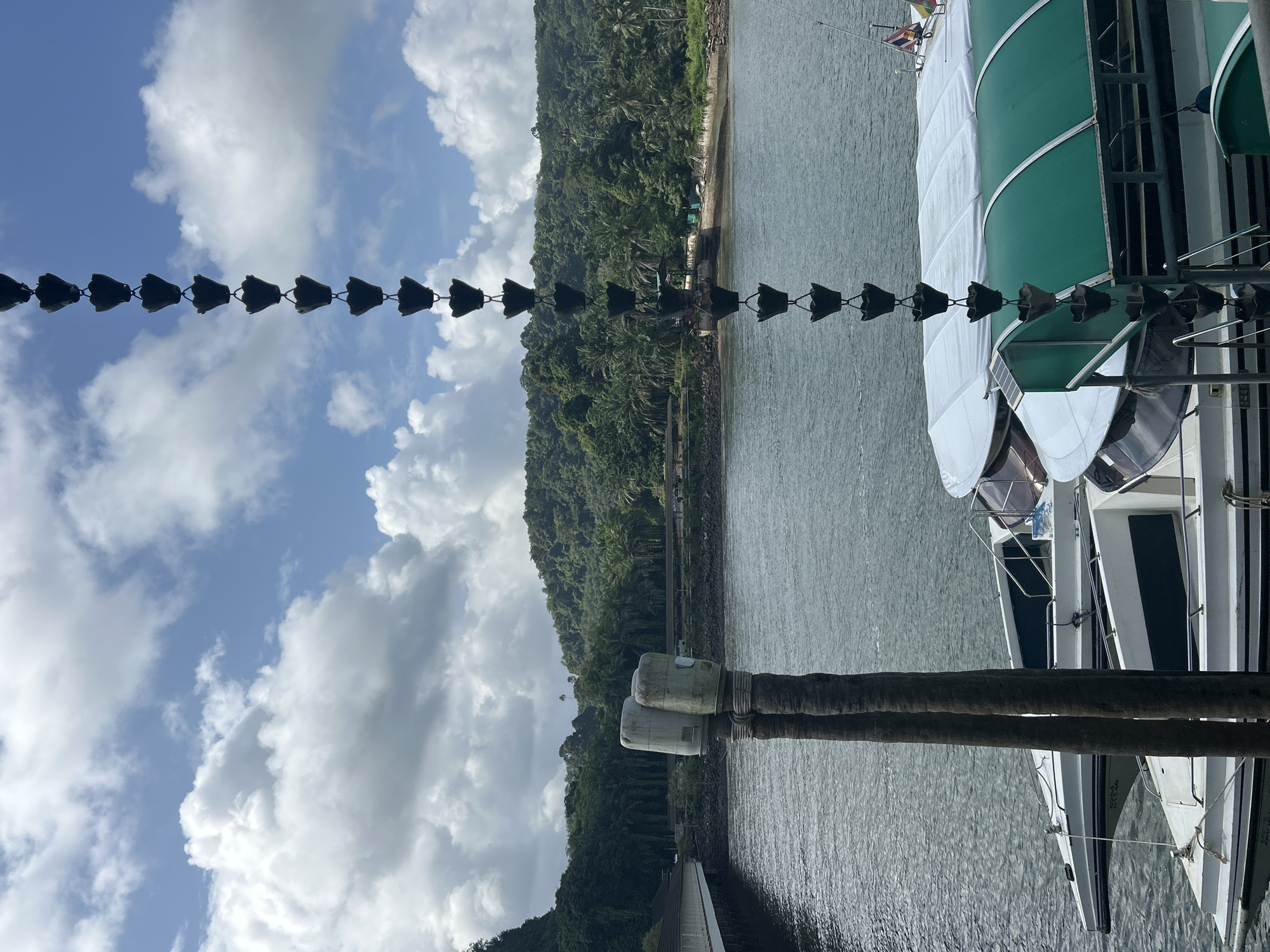 The image depicts a waterfront scene in Pak Nam, Thailand. In the foreground, a decorative metal chain with cup-like segments hangs vertically, obscuring part of the view. Two thick, rough-barked wooden posts, topped with two weathered, cylindrical structures, stand in the water to the left.
Moored in the water are several white boats with dark trim and tinted windows. One boat in the foreground has a white canvas cover, while another boat behind it features a green arched canopy. The water appears calm and slightly rippled.
In the background, a body of water extends towards a lush, green shoreline covered in dense tropical vegetation and palm trees. A low-lying building with a covered walkway or pier is visible on the left side, extending over the water. Further inland, a gently sloping hill is densely forested. The sky above is a mix of blue sky with scattered white cumulus clouds. The overall impression is a serene tropical setting with nautical elements.