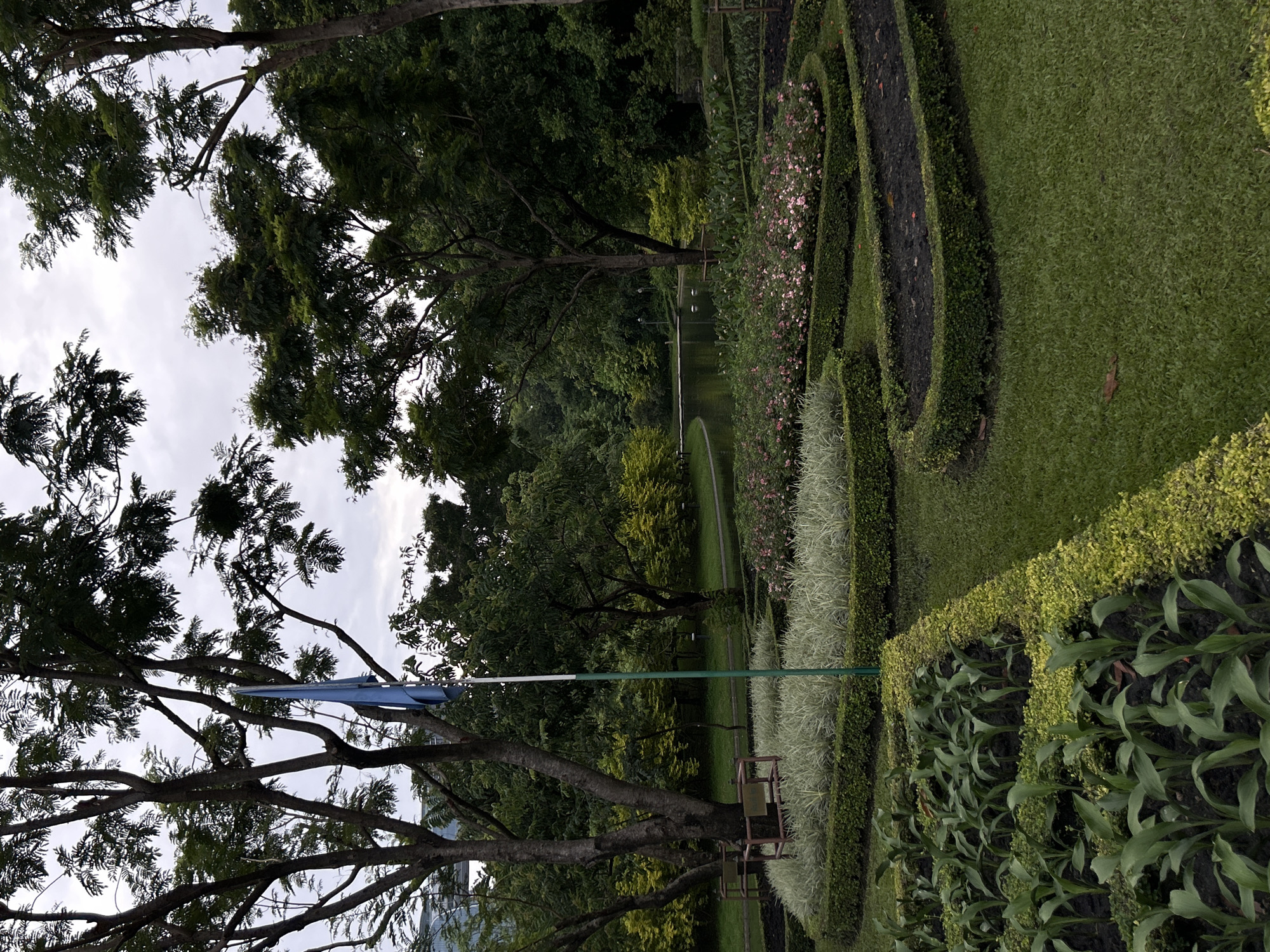 The image depicts a serene park scene with lush greenery and a tranquil pond. In the foreground, neatly trimmed hedges form intricate patterns, surrounding beds of young plants and delicate pink flowers. A tall flagstaff with a blue flag stands prominently near the center. Beyond the flowerbeds, a curved path leads to a serene pond, reflecting the overcast sky and the surrounding trees. Dense foliage and mature trees create a sense of depth and enclosure, with hints of urban structures visible in the distance, suggesting an oasis of nature within the city. This photograph was taken in Lumphini Park, Bangkok, Thailand.