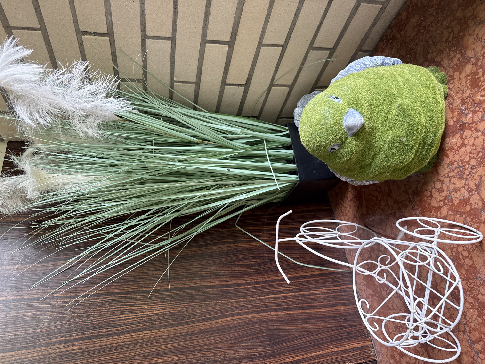 In Herford, Germany, on a reddish-brown speckled surface, a white wire planter shaped like a bicycle sits to the left of a large, green, fuzzy bird figurine. Behind the planter and bird is a large black pot filled with tall, thin, green artificial grass with white fluffy plumes at the top. To the right of the grass is a light brown brick wall. The background to the left of the grass is a dark wood-grained surface.