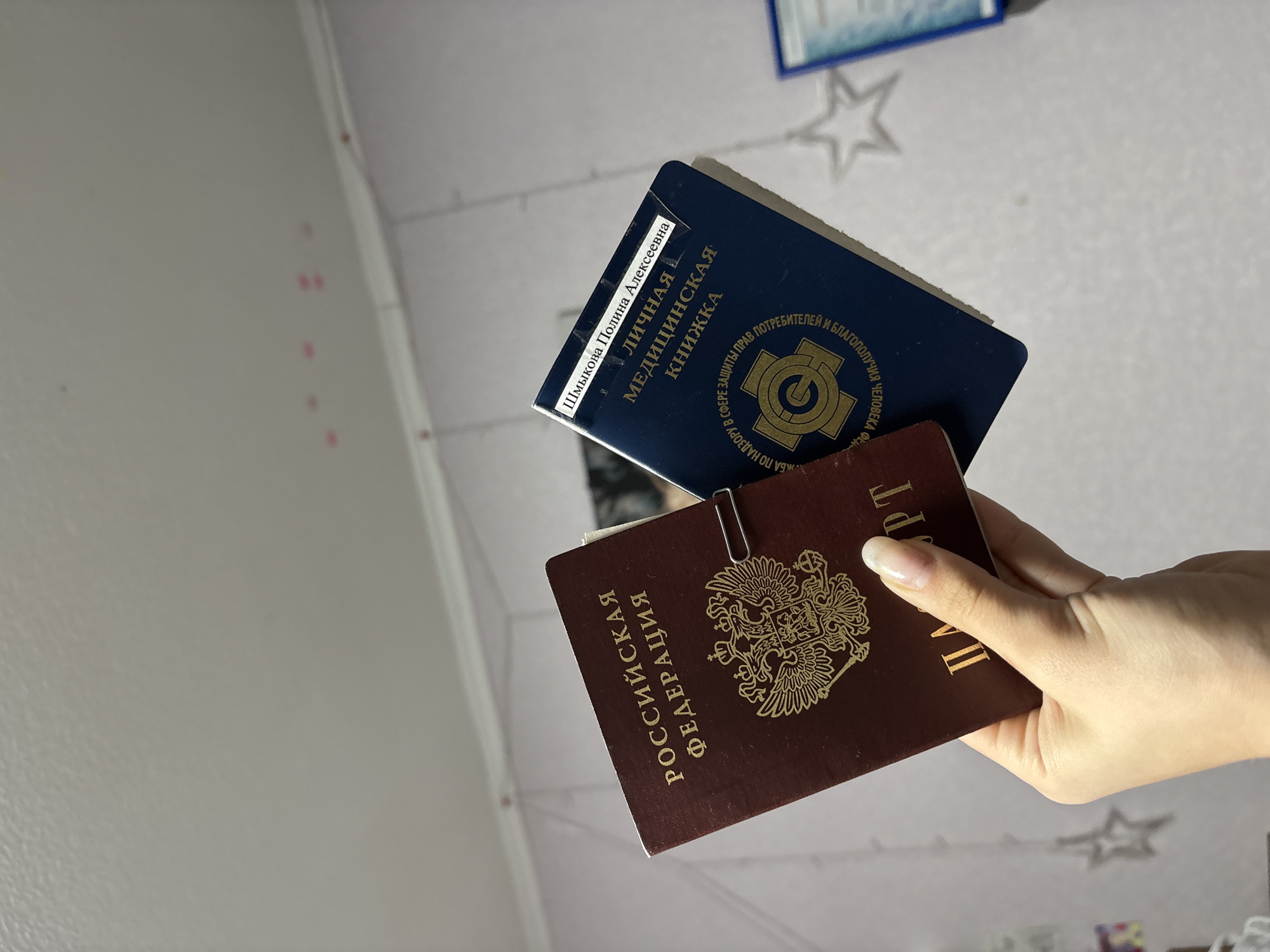 A hand holds two Russian documents against a white wall with decorative stars. The document on the bottom is a maroon Russian passport, secured with a paperclip. Text on the passport reads "РОССИЙСКАЯ ФЕДЕРАЦИЯ" (Russian Federation) and "PT". The top document is a dark blue "ЛИЧНАЯ МЕДИЦИНСКАЯ КНИЖКА" (Personal Medical Book) with the name "Шмыкова Полина Алексеевна". The text "ПО НАДЗОРУ В СФЕРЕ ЗАЩИТЫ ПРАВ ОТРЕБИТЕЛЕЙ И БЛАГОПОЛУЧИЯ ЧЕЛОВЕКА" (for supervision in the field of consumer rights protection and human well-being) is also present. The documents suggest the image was taken in Russia, but there is no specific information to indicate the city of Прикамский.