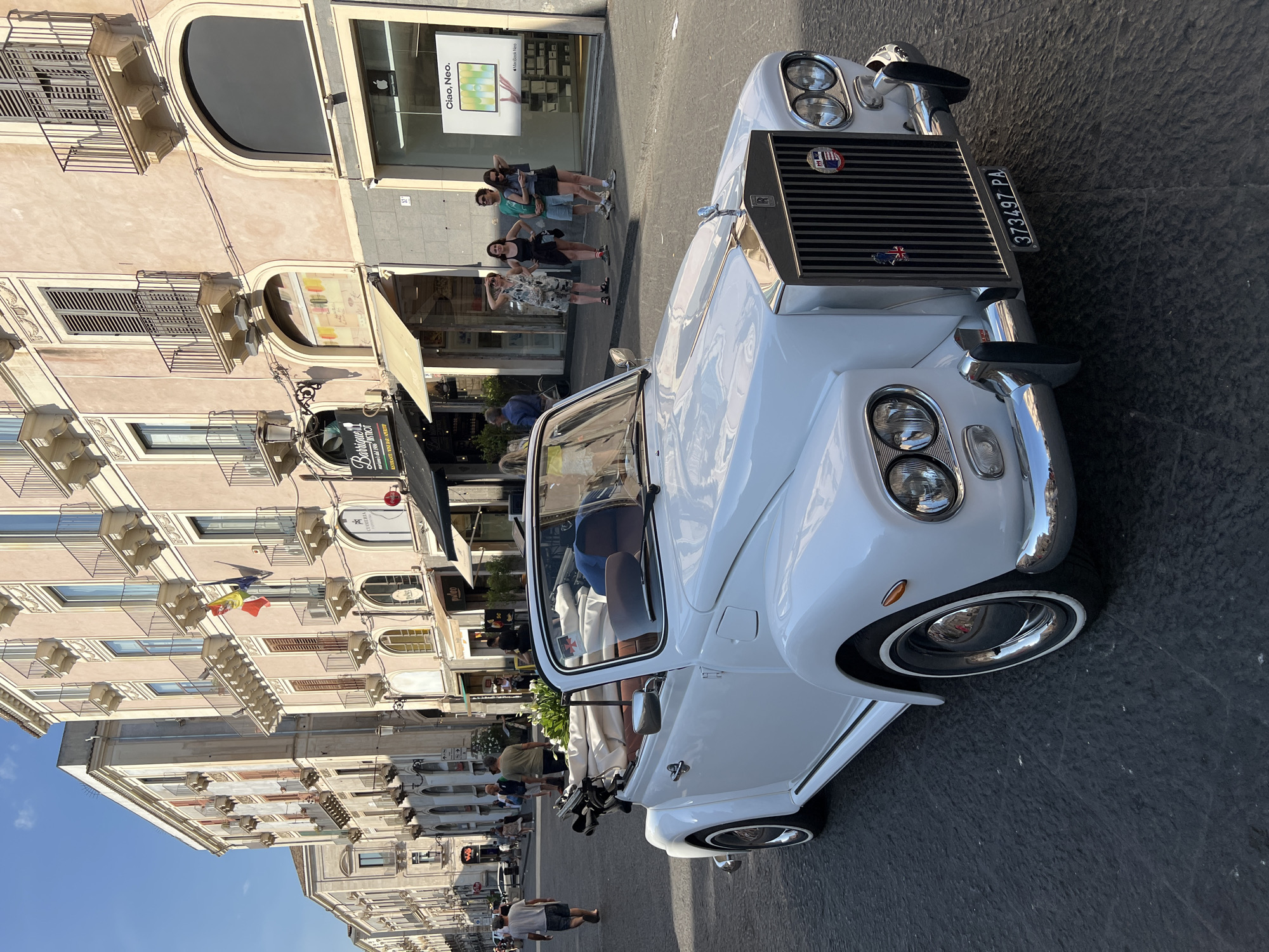 This image captures a street scene in Catania, Italy, likely during daylight hours given the bright sunlight casting sharp shadows. The primary subject is a classic white convertible car, possibly a Rolls-Royce, parked on a cobblestone street. The car features a prominent chrome grille, dual headlights, and a license plate reading "373497 PA".
In the background, a building with multiple stories and balconies lines the street. Shops are visible at street level, one with an awning and another displaying a sign for "Barrique BISTROT ESTORANTE WINE BAR APERITIVO". A window display shows a sign with the text "Ciao, Neo." and an image of a MacBook Neo, suggesting a technology or retail store.
Several people are present in the scene. Four women are standing together in front of the store window, appearing to be posing for a photo or simply socializing. Further down the street, other pedestrians can be seen walking. The overall atmosphere seems relaxed and casual, typical of a sunny day in a European city. The presence of Italian text on the signs confirms the location as Italy, specifically Catania as per the provided context. The buildings exhibit architectural details common in Italian cities, with ornate balconies and stucco facades. The weather appears clear and sunny.