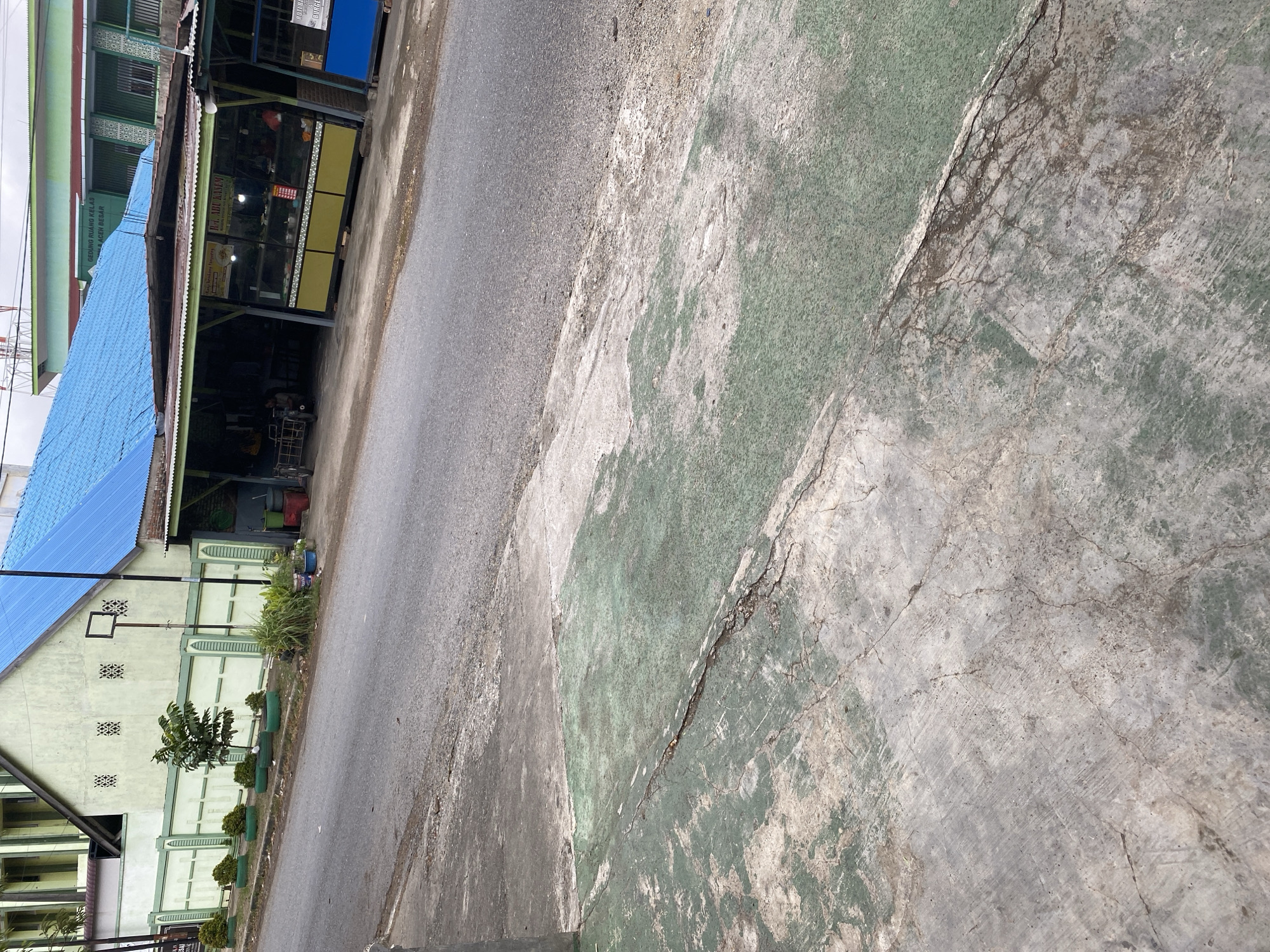 The image depicts a street scene in Payatieng, Indonesia. In the foreground, a cracked and worn concrete surface with patches of faded green paint dominates the lower portion of the frame. The surface transitions into an asphalt road, suggesting a public thoroughfare.
In the mid-ground, a light green building with a blue roof is visible. This building appears to be a school or institutional facility, as indicated by the signage that reads "GEDUNG RUANG KELAS" and "ACEH BESAR." Adjacent to the building, a series of small shops or food stalls with open fronts are set up. One stall, labeled "R.T. ADU KASEM," displays goods in a glass-fronted cabinet. A tricycle is parked near one of the stalls. A short green fence with decorative posts lines the front of the building, with several small, manicured bushes and a young tree planted along its length.
The background shows more of the institutional building and parts of other structures, including what appears to be a water tower or communication antenna. The overall impression is of a typical urban or semi-urban environment in Indonesia, with a mix of residential, commercial, and institutional buildings. The lighting suggests it is daytime, likely overcast or late afternoon. No people are clearly visible or engaged in any specific activities.