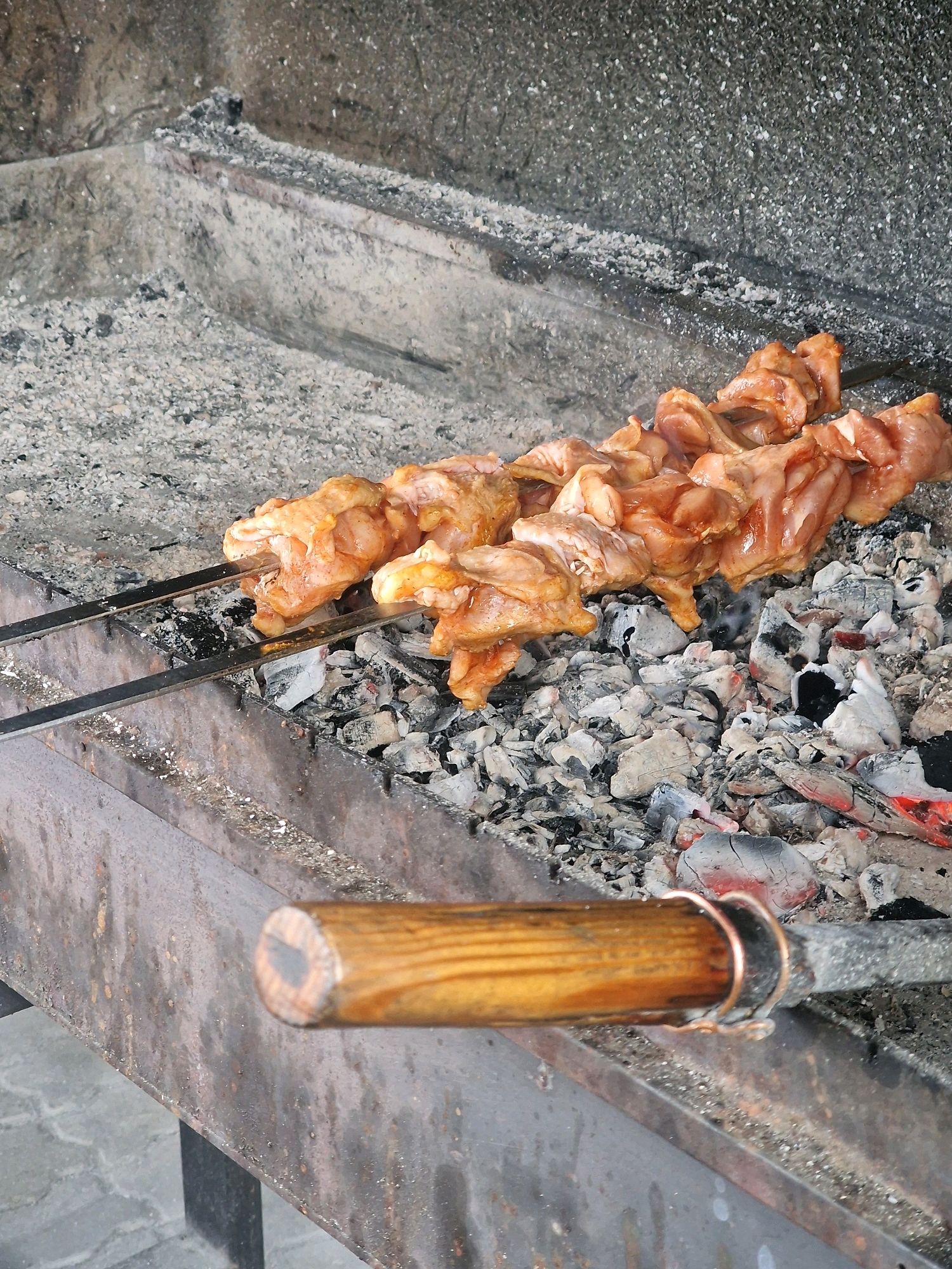 The image shows a close-up view of chicken skewers cooking over hot coals on a grill. The skewers are positioned horizontally, with raw chicken pieces threaded onto metal rods. The chicken appears to be marinated in an orange-colored sauce. The grill itself is made of metal and is filled with ash and glowing red embers. A wooden handle of a tool, possibly a grill brush or skewer holder, is visible in the foreground. The background shows the rough, textured surface of the grill's interior, which is covered in ash. This scene is consistent with outdoor cooking, and could be found in a variety of locations, including a backyard, a park, or a street food vendor setup in Zelenogradsk, Russia, given the context provided.