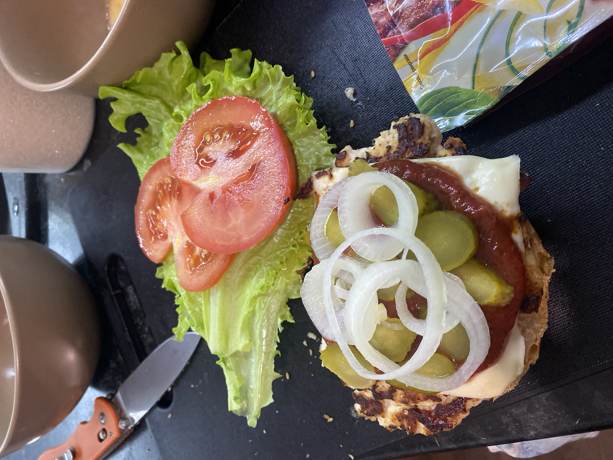 The image shows an open-faced sandwich or burger on a black cutting board. The ingredients include lettuce, sliced tomatoes, a cooked patty with melted cheese, ketchup or a similar red sauce, pickles, and sliced onions. A pocketknife with an orange handle is lying near the ingredients. Bowls are partially visible in the upper left. In the upper right corner, there's a blurry package that appears to contain food. No people are visible. There's no information in the image to indicate the location is Izhevsk, Russia.