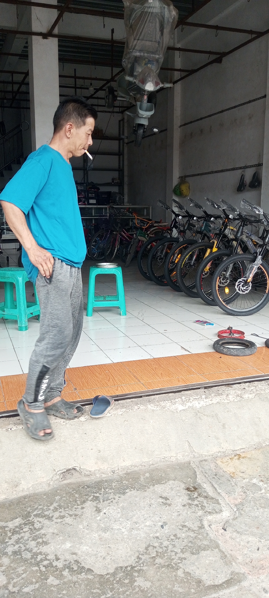 A man is smoking a cigarette while standing in front of a bicycle shop. He is wearing a blue t-shirt and grey sweatpants. There are several bicycles lined up in the background. A scooter is hanging from the ceiling, covered in plastic. The shop appears to be located in Setu, Indonesia. The ground is tiled inside and concrete outside the shop.