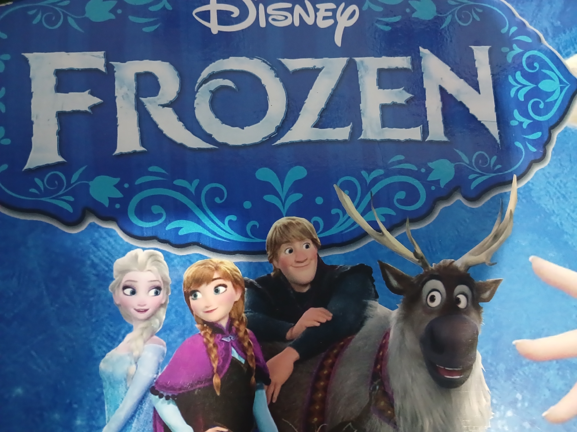 The image depicts characters from the Disney movie "Frozen" on a blue background with ornate silver and turquoise designs. In the foreground, a reindeer with large eyes and antlers is partially visible. Behind the reindeer, a man with blond hair and a black shirt is seated. Anna, with brown braided hair and a purple cape, is behind him. Elsa, with blond braided hair and a light blue dress, is in the background. The word "FROZEN" is written vertically in large white letters on the right side of the image, with the Disney logo also present. The image is likely a poster or merchandise associated with the movie. There is no information in the image to determine its location in Randuagung, Indonesia.