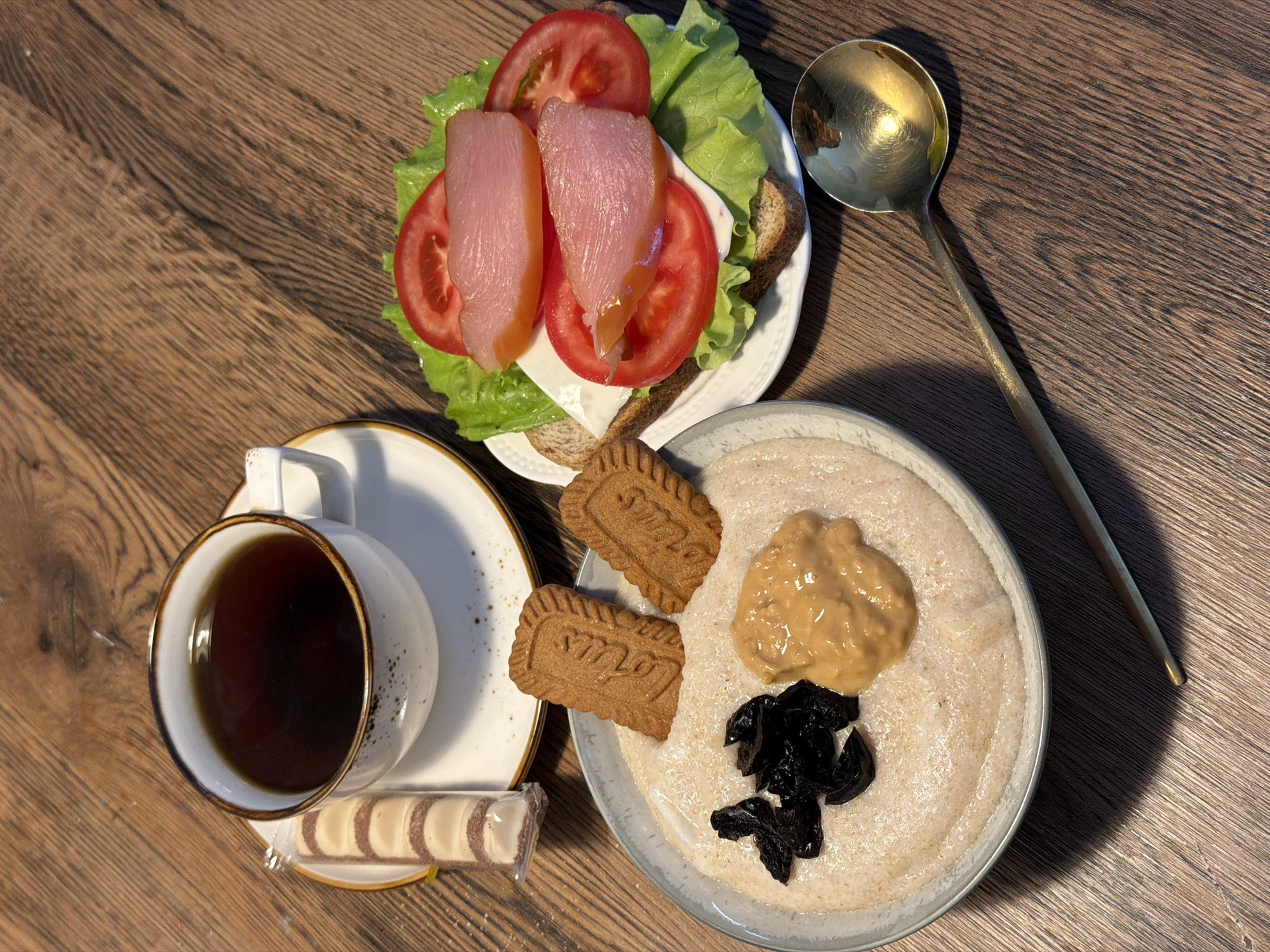 A high-angle medium shot shows a breakfast spread on a wooden table. The meal includes a bowl of oatmeal topped with prunes and peanut butter, two Lotus Biscoff cookies, a sandwich with lettuce, tomato, cheese, and meat, a cup of coffee with a wrapper of white chocolate wafer snack, and a golden spoon. The scene is likely indoors, as indicated by the lighting and table surface, and no specific location in the city of Perm, Russia is discernable.