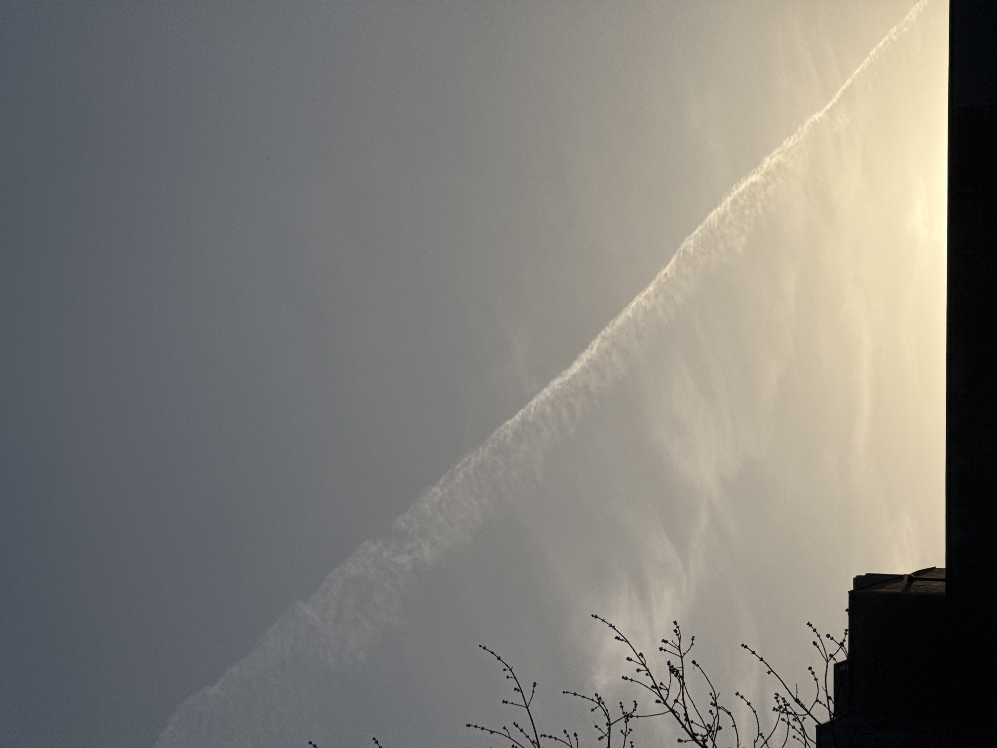 The image depicts a sky with a diagonal contrail stretching from the bottom left to the upper right. The sky transitions from darker grey on the left to a brighter, yellowish-grey on the right, where the sun appears to be located. A silhouette of a building corner is visible on the right side of the image. In the lower part of the picture, bare branches of a tree are visible against the sky.
I can not say that this picture taken in the city of Yekaterinburg, Russia, because I have no way to check this fact.
