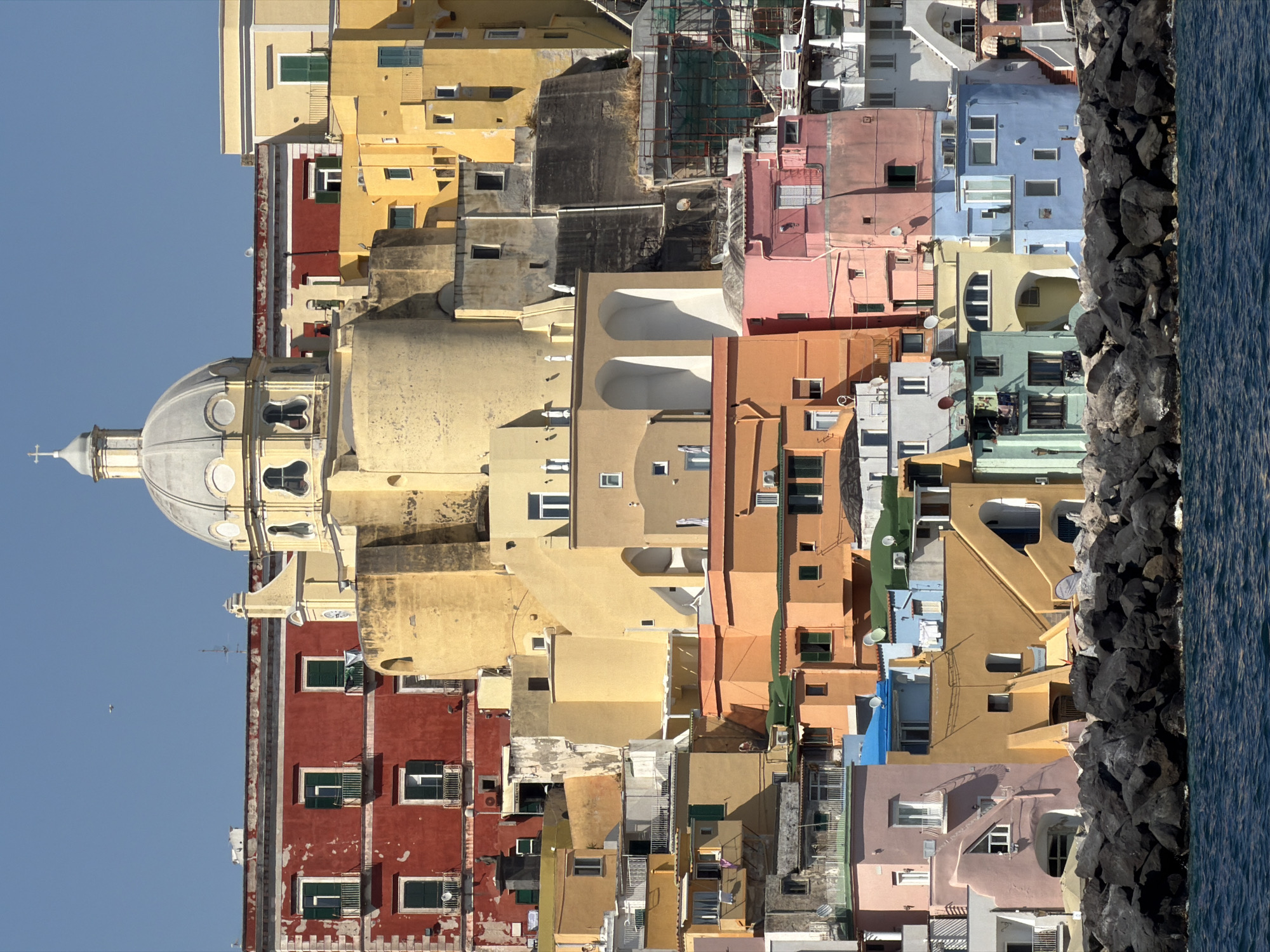An upward-facing outdoor perspective showcases a dense cluster of buildings against a clear, light blue sky on the left and the dark blue sea separated by dark rocks on the right. The structures exhibit a diverse range of colors, including yellows, reds, pinks, blues, and greens. The facades of the buildings feature numerous windows and architectural details. Dominating the image is the dome of the church. This appears to be the colorful architecture of Procida, Italy.