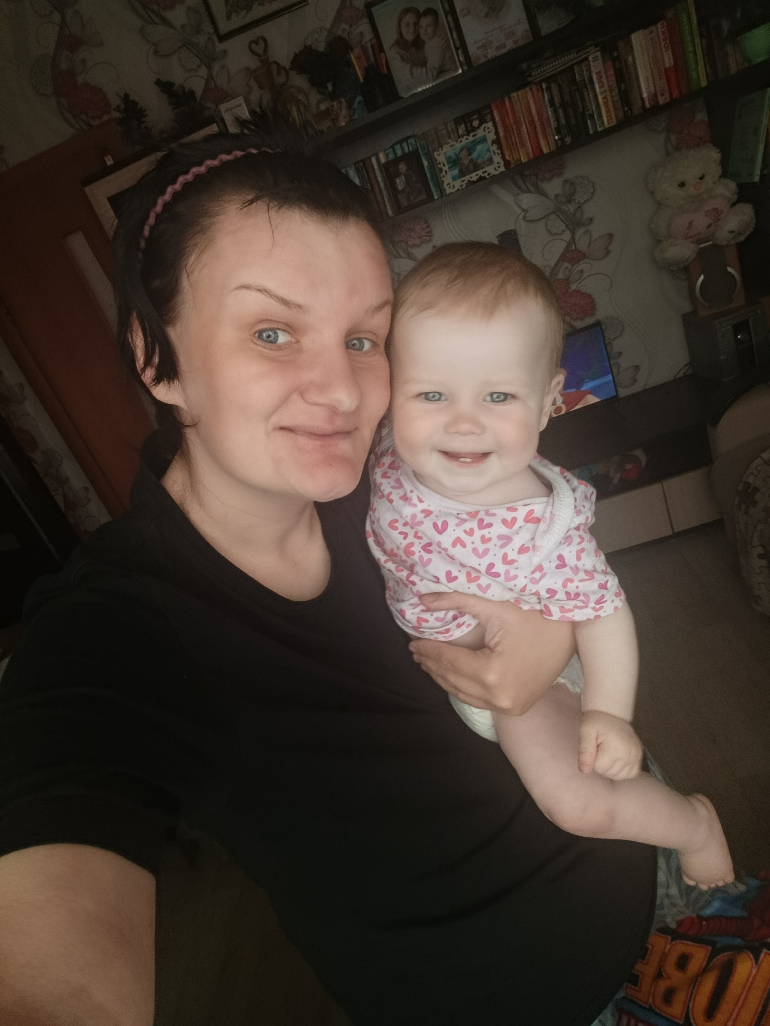 A woman with short, dark hair and blue eyes is holding a baby with light hair and blue eyes. The woman is wearing a black shirt and a thin headband. The baby is wearing a white onesie with a pink heart pattern and a diaper. They are both smiling.
Behind them is a wall covered in a floral wallpaper. There is a dark shelf with books and framed pictures on the wall. A white teddy bear with a heart on it is also visible on the shelf. A television is visible on a console under the shelves. There are objects beneath the television and on the floor to the bottom right of the image.
While the city is mentioned as Фоки, Russia, there is no detail visible in the image to confirm this location.