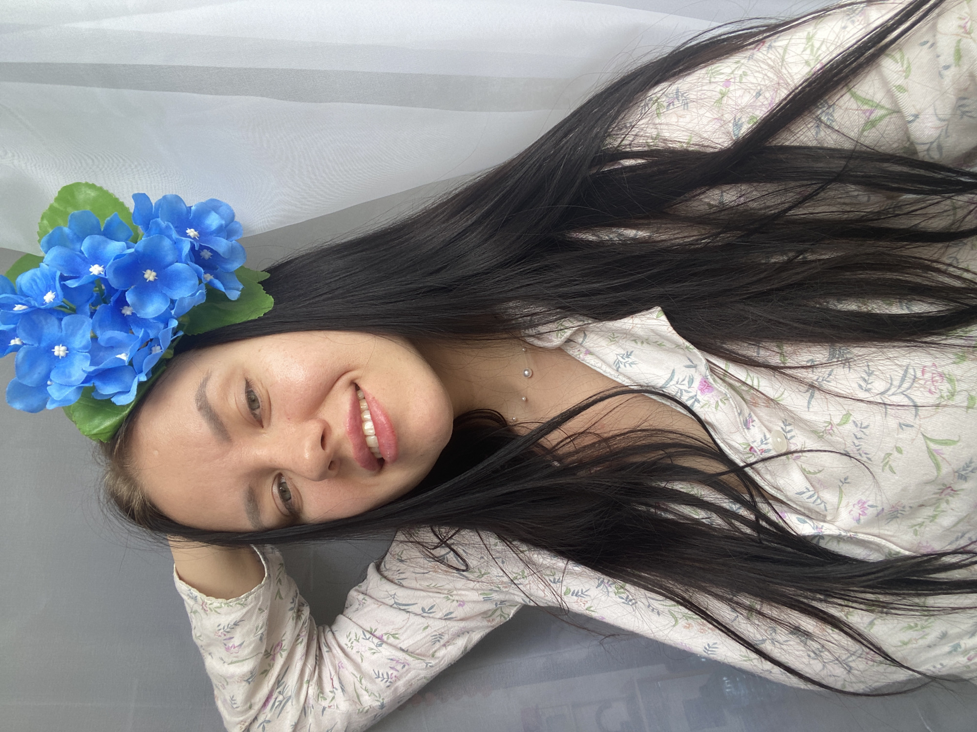 A young woman with long, dark hair and blue flowers in her hair is posing for a selfie. She is wearing a light-colored, floral-patterned shirt. She is smiling at the camera with her left hand behind her head. The background is a gray curtain on the left and a white sheer curtain on the right. The image appears to be taken indoors. The location is not specified in the image or provided in the prompt.
