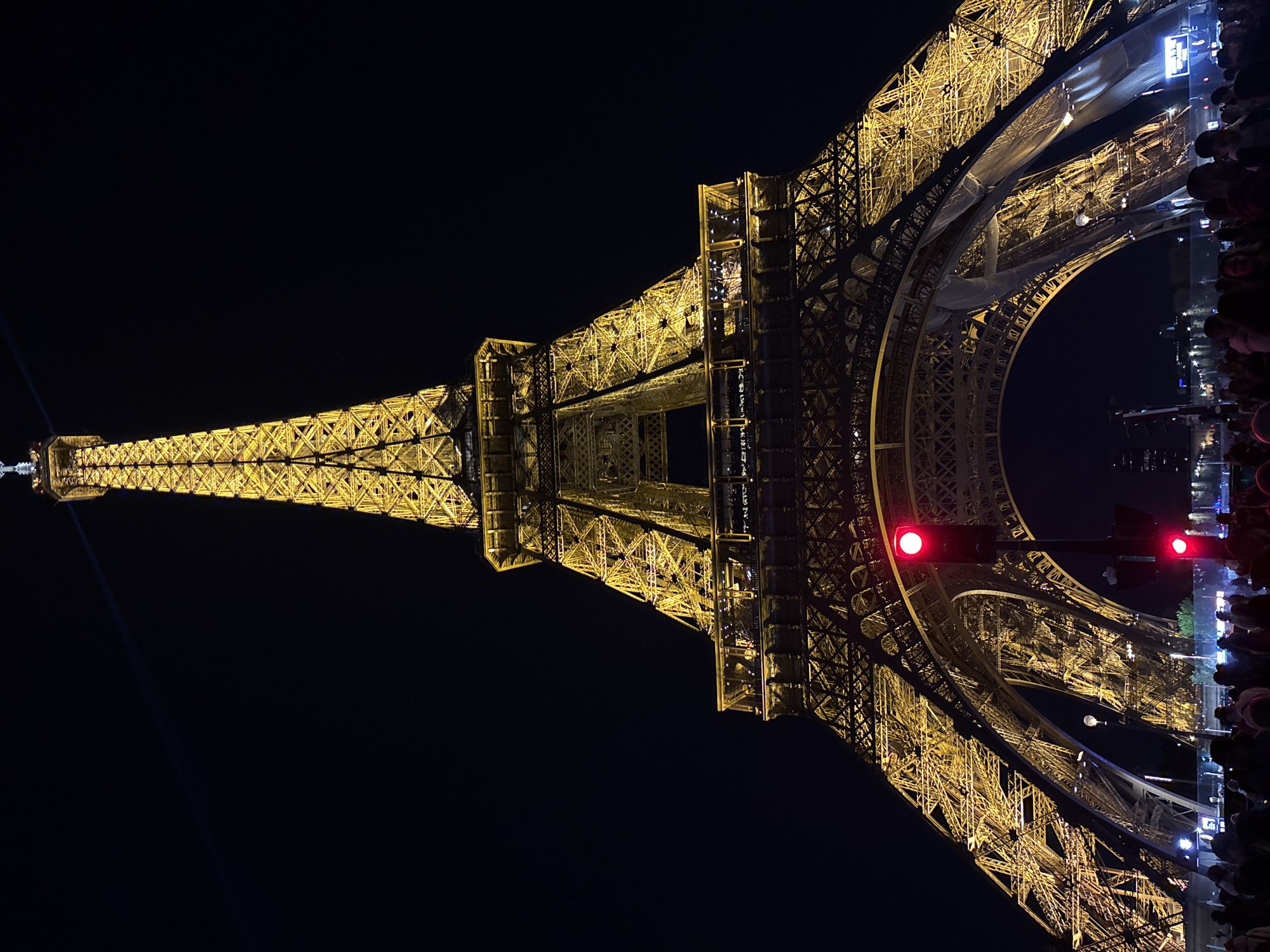 At night, the illuminated Eiffel Tower in Paris, France, is visible, with a dark sky in the background. The tower's metal structure is highlighted with yellow lights. Red traffic lights are also visible near the base of the tower. A crowd of people is gathered to the right of the tower. A beam of light is emitted from the tower's top.