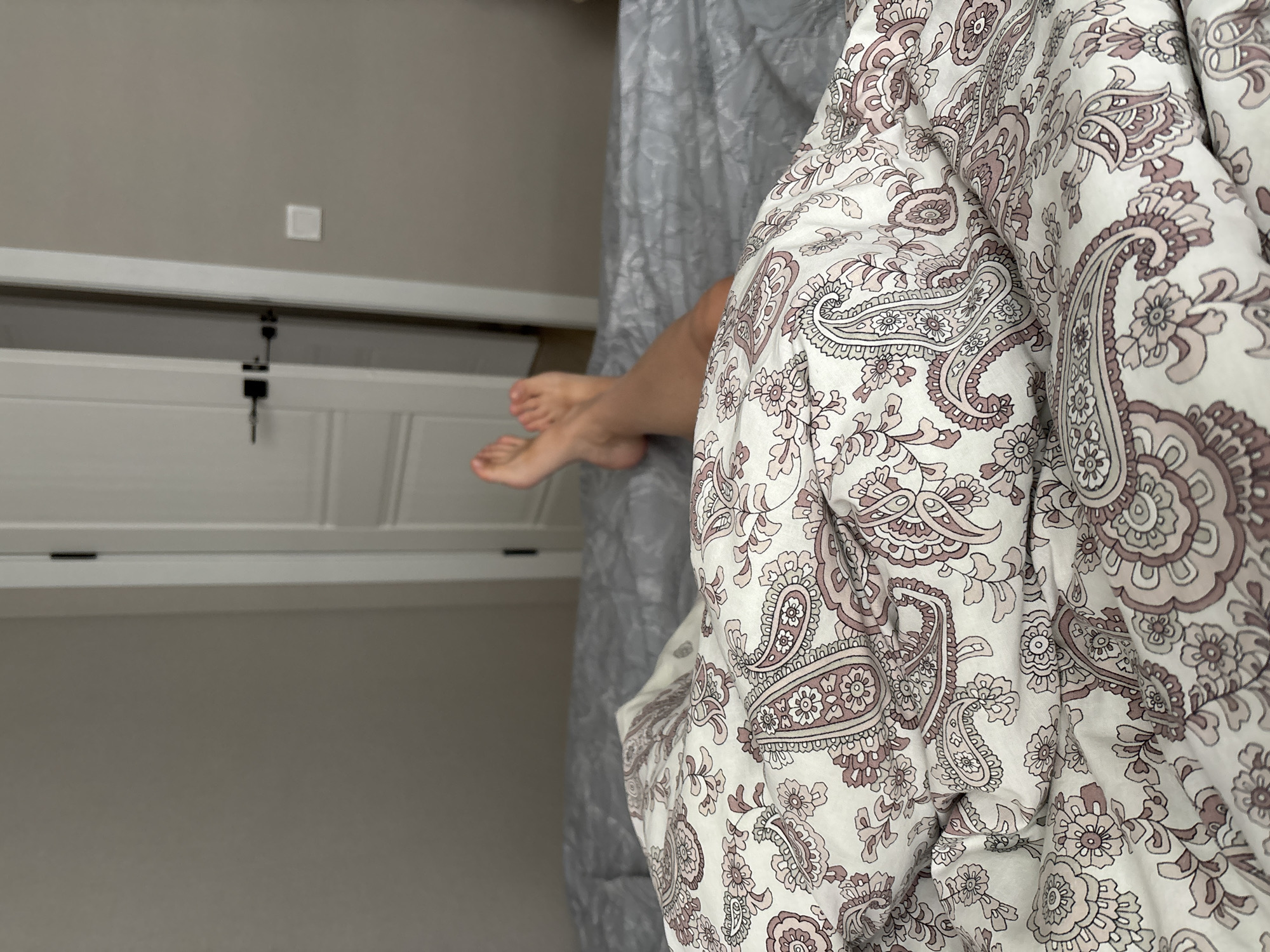 The image shows a close-up view of a person's feet and legs resting on a bed. The bed is covered with a duvet featuring a paisley pattern in shades of pink and white, and a gray comforter underneath. The background shows a doorway with a white door and dark handle, and a neutral-colored wall. The scene is indoors, likely in a bedroom. There are no identifiable landmarks or contextual clues to determine the specific location within the city of Chaikovsky, Russia.
