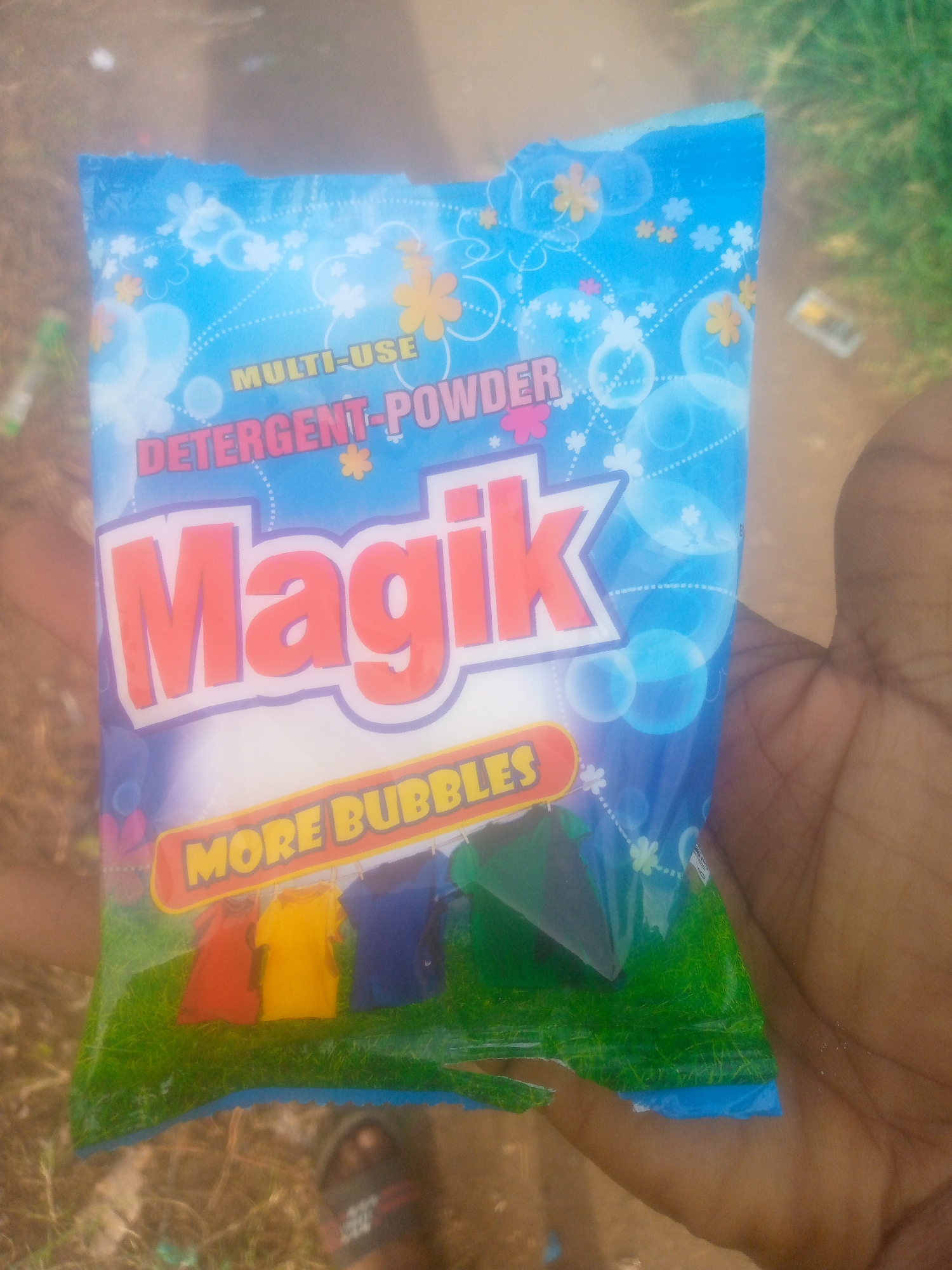 A hand is holding a packet of "Magik" multi-use detergent powder. The packet has a blue background with floral and bubble designs. The word "Magik" is written in large, red letters. Below, there is an image of clothes hanging on a line over a green field. The photo appears to be taken outdoors, with grass and other debris visible in the background. A foot wearing a sandal is partially visible in the lower portion of the image. The location is unspecified.