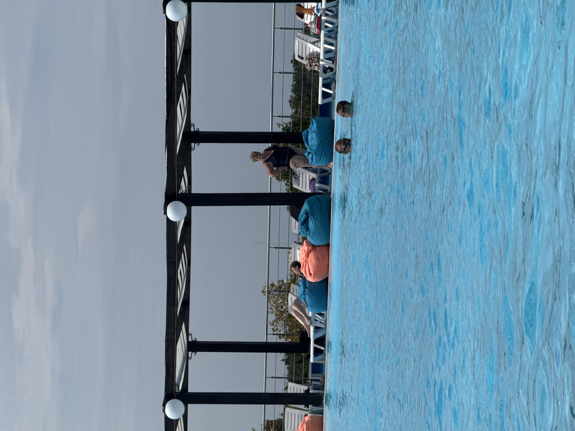 An outdoor swimming pool scene is depicted. In the foreground, the blue water of the pool is disturbed by ripples. Two people, a man and a child, are visible in the water, both wearing goggles. In the mid-ground, a woman in a dark swimsuit stands near several large, beanbag-style chairs. Behind her, other people are relaxing on lounge chairs. A shaded pergola structure with white spherical lights is visible across the top of the mid-ground. The background consists of a railing, trees, and a cloudy sky. The location is identified as Sevastopol, Ukraine.