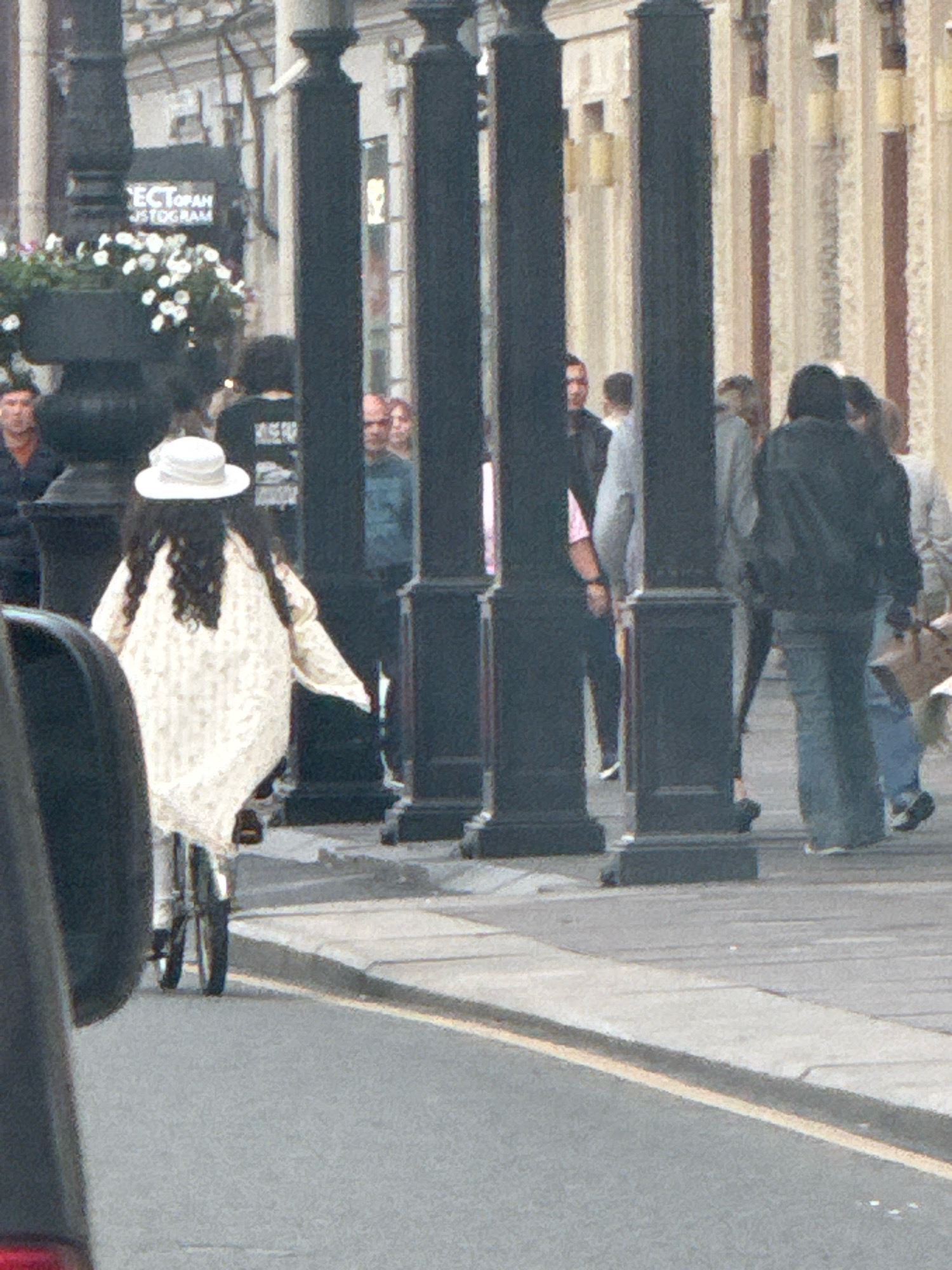A group of people are walking and standing on a sidewalk and street. In the foreground, a person is riding a bicycle. The person is wearing a white hat and a cream-colored patterned cloak. The background shows a building with tall columns and windows. There are also several black posts on the sidewalk. The scene appears to be in a city, possibly Saint Petersburg, Russia, given the architectural style. The lighting suggests it is daytime.