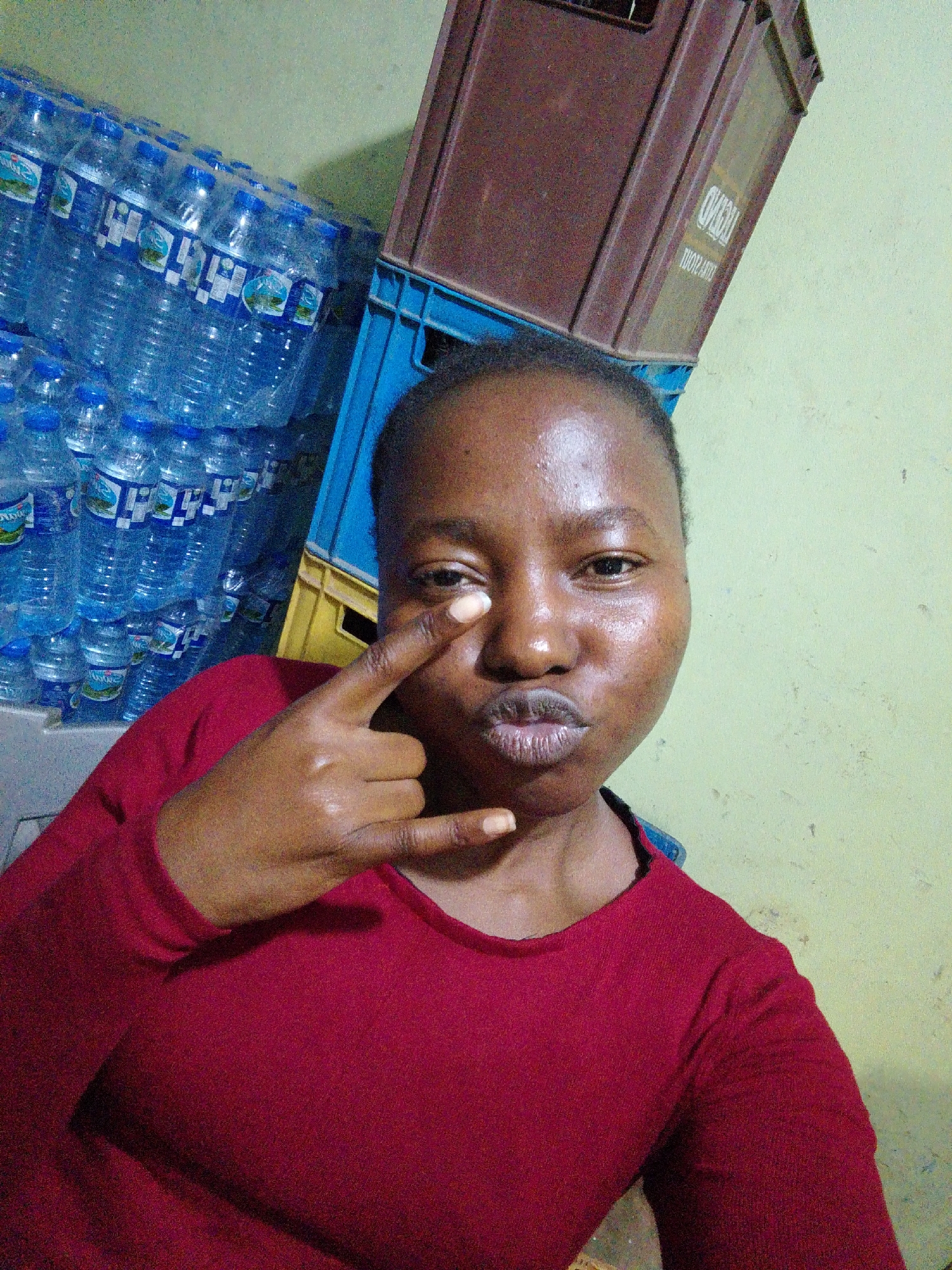 The image is a selfie of a dark-skinned person wearing a long-sleeved red shirt. They are making a gesture with their right hand, holding their index and pinky fingers up near their eye. The background consists of stacks of bottled water and two crates. The location is likely indoors in an unknown area of the city of Unknown, Unknown. The overall lighting is dim and slightly yellow.
