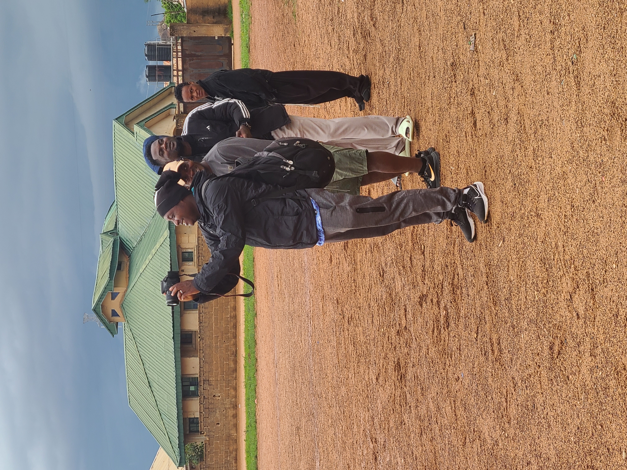 An outdoor image, presented on its side, shows four men standing in a line on a reddish-brown, gravelly ground. The men are dressed in casual, athletic-style clothing.
The man closest to the viewer is positioned on the left and appears to be holding a black digital camera with both hands, seemingly taking a picture or recording. He wears a black jacket, grey pants with blue detailing on the side, and a black beanie. He has black and white sneakers.
Behind him, a man with a beard is visible, wearing a grey shirt, olive green shorts, and a black backpack. His shoes are light green.
Further back, a third man wears a dark blue beanie, a black Adidas jacket with white stripes on the sleeves, light-colored pants, and light-colored sneakers.
The last man in the line, furthest from the camera, is wearing a black tracksuit.
In the background, on the left side of the frame, there is a building with a distinctive green-tiled roof and light-colored walls. On the right side of the background, two large, black cylindrical water tanks are visible on a raised platform, with a brick wall behind them.
The sky overhead is clear and light blue, indicating daytime, likely under bright conditions. The ground beneath the men is a uniform texture of small, reddish-brown pebbles or earth, suggesting an unpaved outdoor area like a field or yard. No visible text is discernible on clothing or objects. The atmosphere appears casual and relaxed.