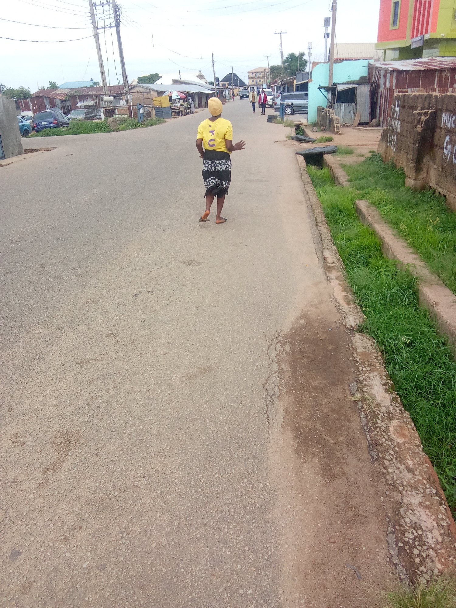 In the city of Jos, Nigeria, a woman wearing a yellow shirt and a patterned skirt is walking on a paved road. She is facing away from the camera. The road is lined with buildings, some appearing to be shops or residences. There are also some vehicles on the road, including a blue car and a van. The environment appears to be a typical urban street scene with signs of commercial activity and residential housing. The sky is overcast.