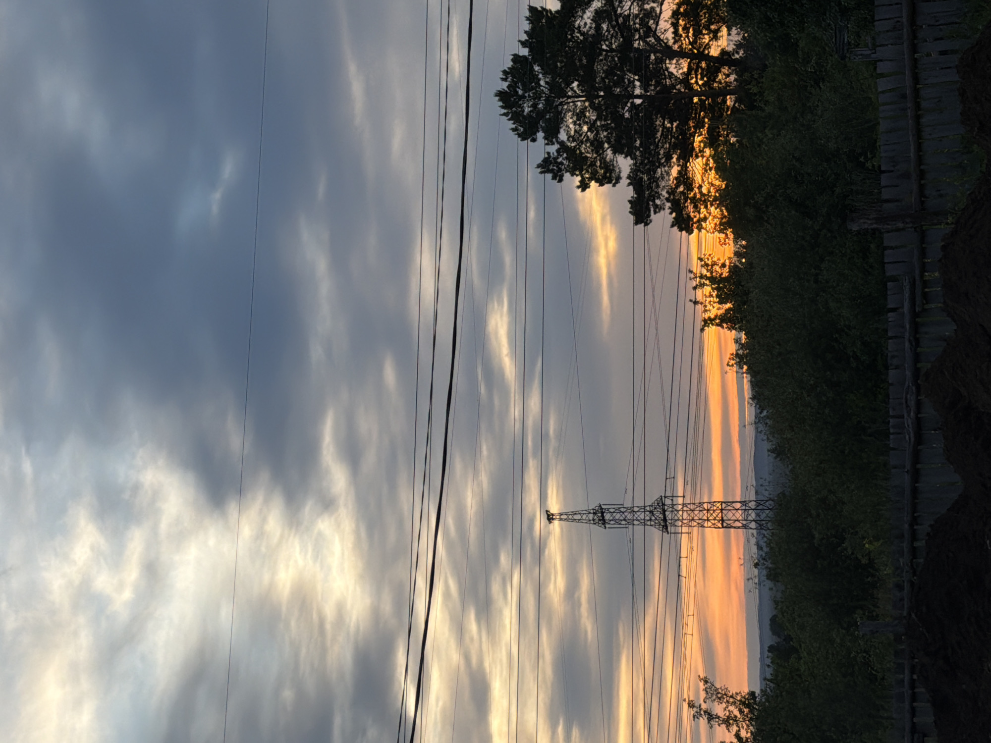 The image captures a sunset in Chaikovsky, Russia, evidenced by the golden light illuminating the sky. High-voltage power lines stretch across the frame, supported by a lattice tower. The foreground includes dense greenery and a wooden fence. The sky is filled with textured clouds, ranging from dark grey to light yellow where the sun is setting.