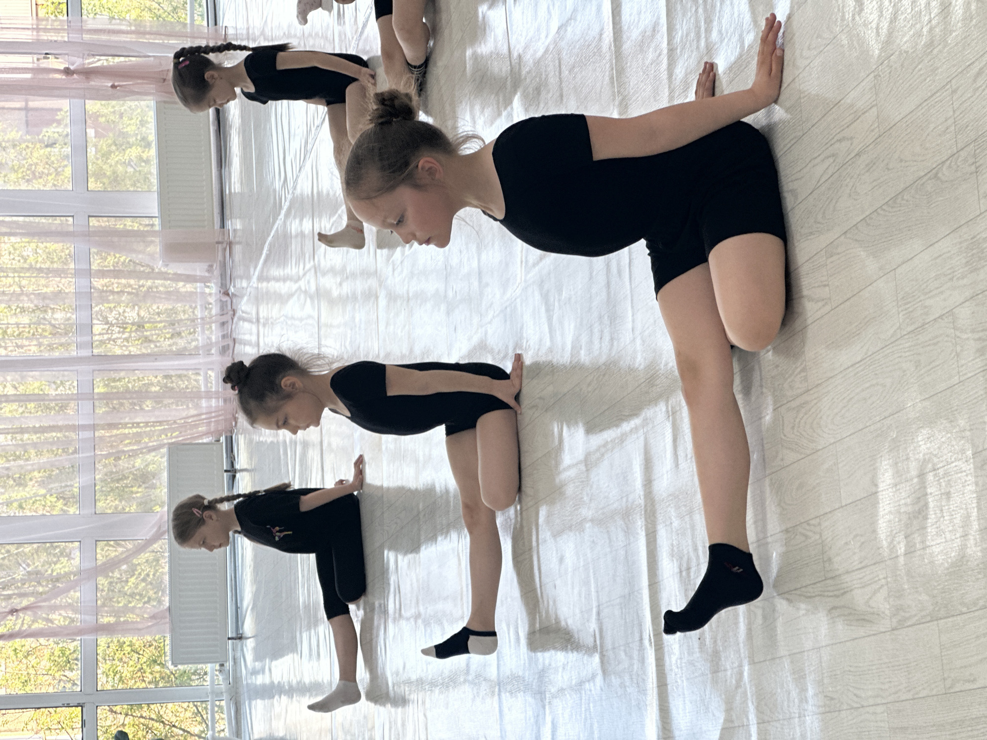 The image depicts four young girls in a dance studio or similar space in Votkinsk, Russia. They are positioned in a line, each performing a stretching exercise against what appears to be a wall covered with a white cloth or backdrop.
The girls are dressed in black leotards. The tallest girl in the foreground wears black socks on one foot. They each have their hands pressed against the wall at shoulder height and are bending forward at the waist.
The room has large windows with sheer curtains, allowing natural light. A white radiator is visible under one of the windows. The floor is a light color with a wood grain pattern.