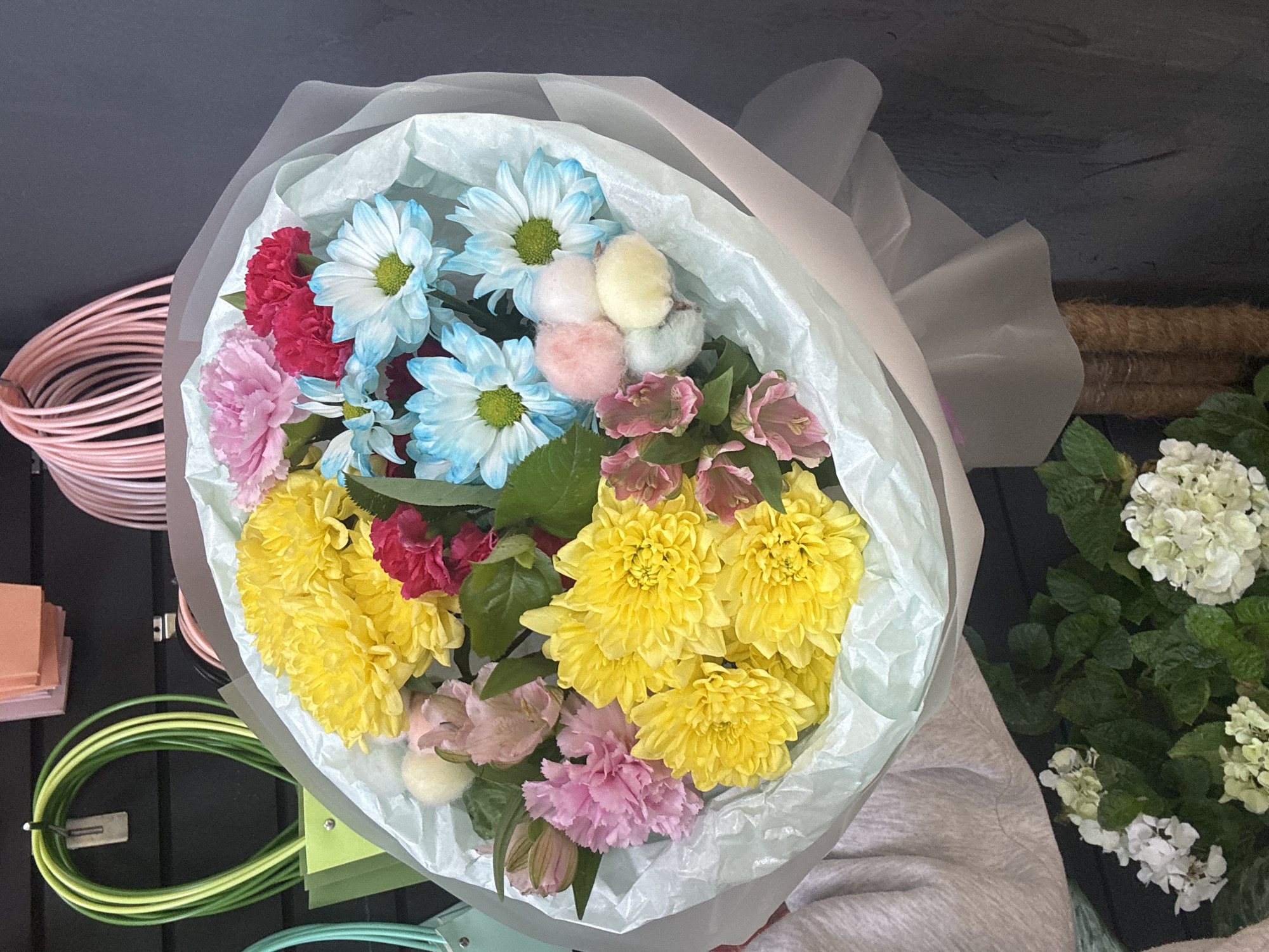 A medium shot captures a vibrant bouquet of flowers, predominantly yellow chrysanthemums, interspersed with delicate pink carnations, bold red carnations, and cheerful blue-tipped daisies. The bouquet is artfully wrapped in pale green and pinkish-white tissue paper, creating a soft, ethereal presentation. A person's arm and shoulder are visible on the left side, suggesting they are holding or presenting the bouquet. In the background, several colorful tote bags with looped handles are displayed against a dark wall. Below the bouquet, a cluster of white hydrangeas adds a contrasting texture and color. The overall setting appears to be a flower shop or a similar retail environment. The lighting is bright and even, highlighting the textures and colors of the flowers and merchandise.