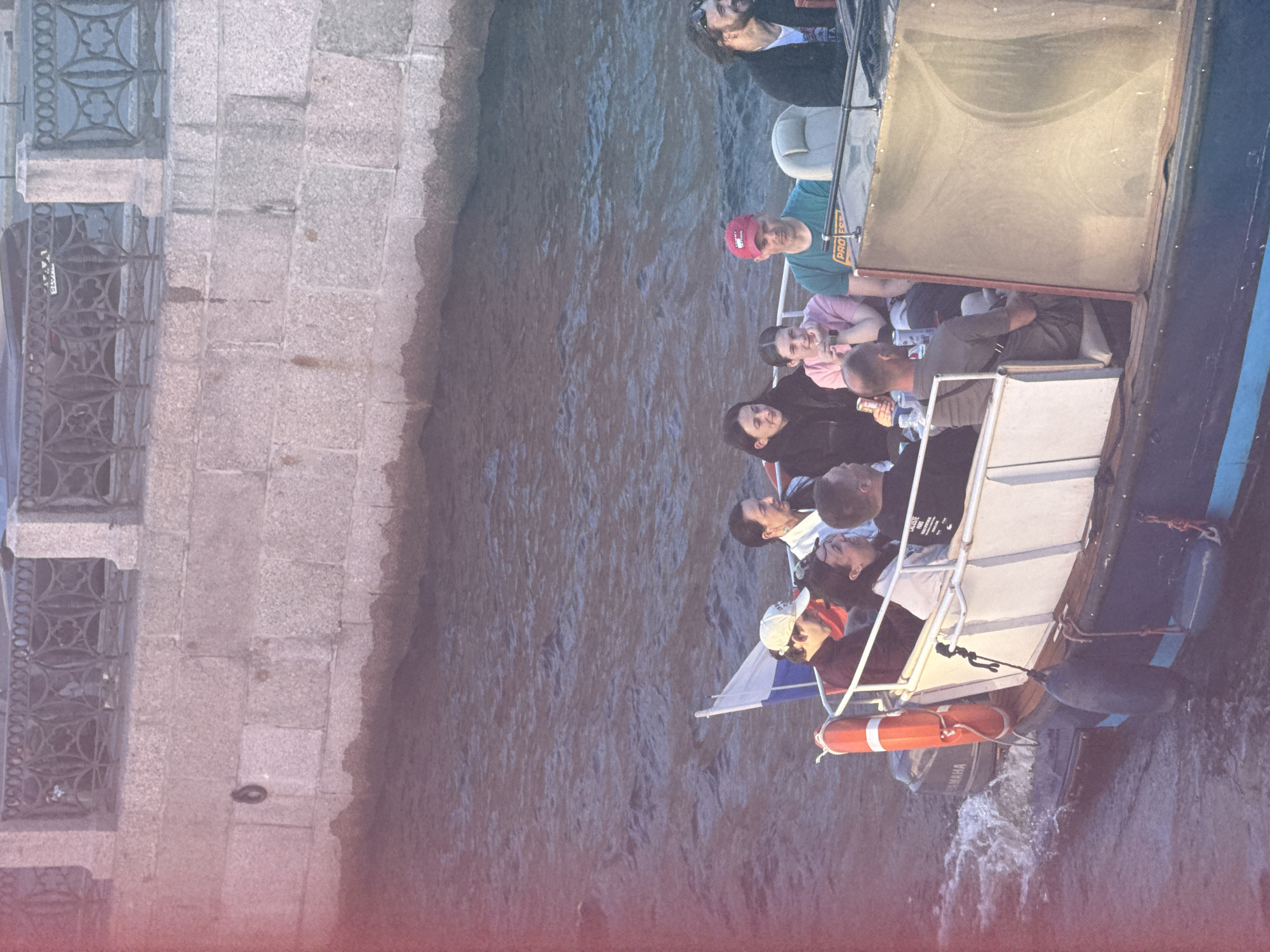 The image depicts a small tour boat on a body of water, likely a canal or river, in Saint Petersburg, Russia. The boat is filled with passengers, many of whom are looking towards the side or at each other. One person is holding a can of soda. The boat is moving, indicated by the wake behind it. In the background, there is a stone embankment with an ornate metal railing. The overall scene suggests a leisurely boat tour in a city known for its waterways and historical architecture. The lighting suggests it is late afternoon or early evening.